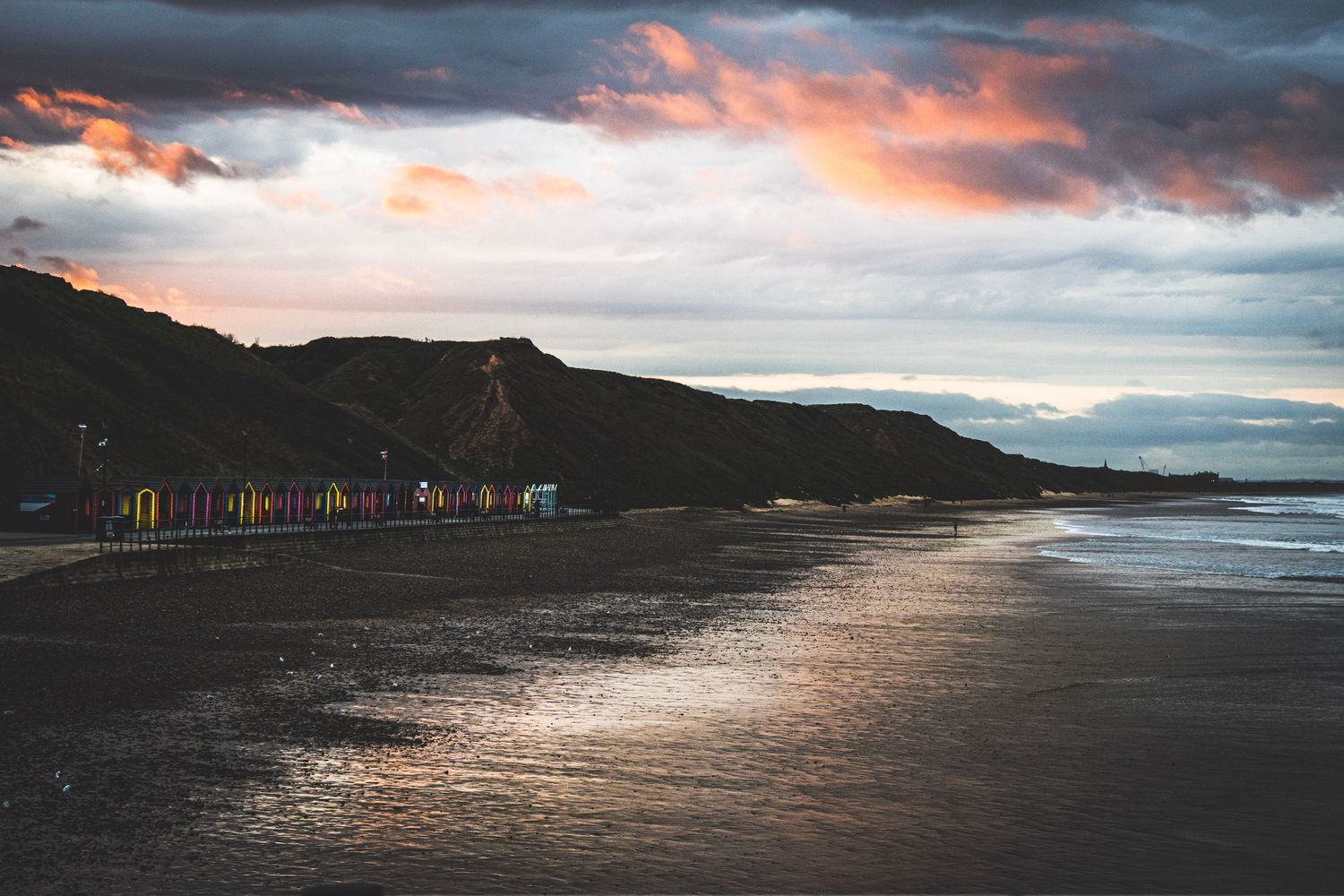 Dramatic sunset over a coastal beach with dark clouds and glowing orange sky reflecting on the ocean water.