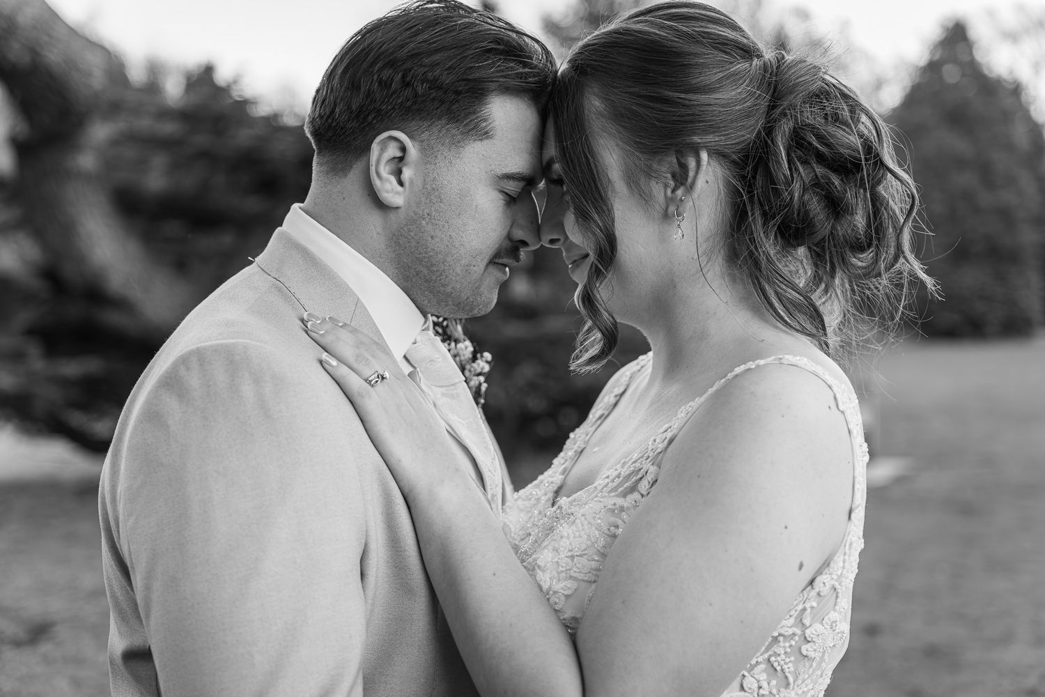 Black and white wedding photo of bride and groom touching foreheads tenderly outdoors.