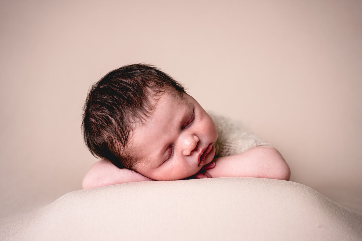 Peaceful newborn baby sleeping on a soft pink pillow with dark hair and tiny hands tucked under chin.