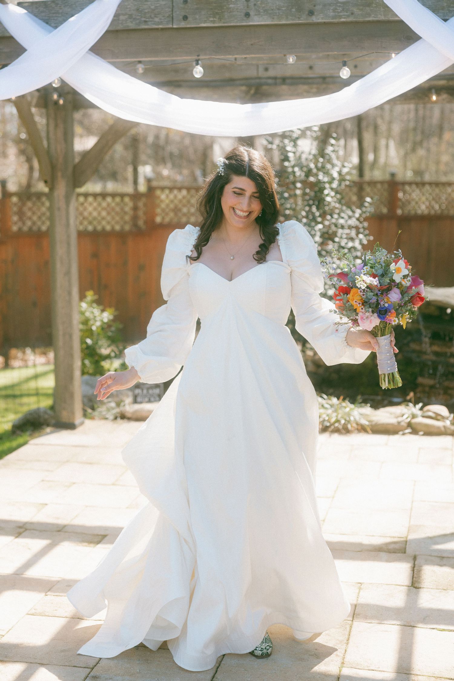 Radiant bride in white long-sleeve gown with puff sleeves holds colorful bouquet under floral arch at outdoor wedding.
