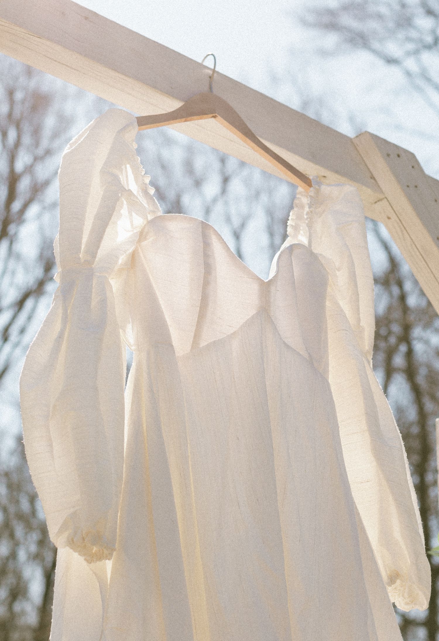 White linen puff-sleeve wedding dress hanging on wooden hanger outdoors against a blue winter sky.
