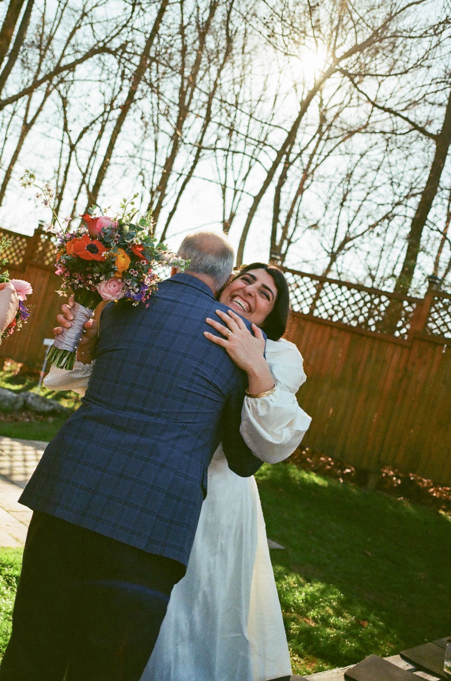 Bride in white dress hugging a man in blue plaid suit at outdoor backyard wedding ceremony with colorful bouquet.
