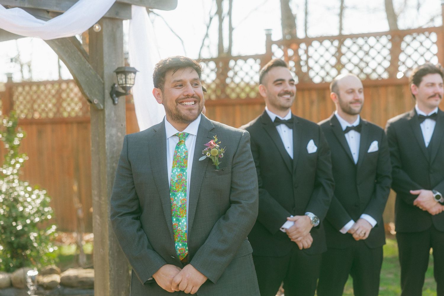 Smiling groom in gray suit with colorful tie stands at altar with groomsmen in black tuxedos at outdoor wedding ceremony.