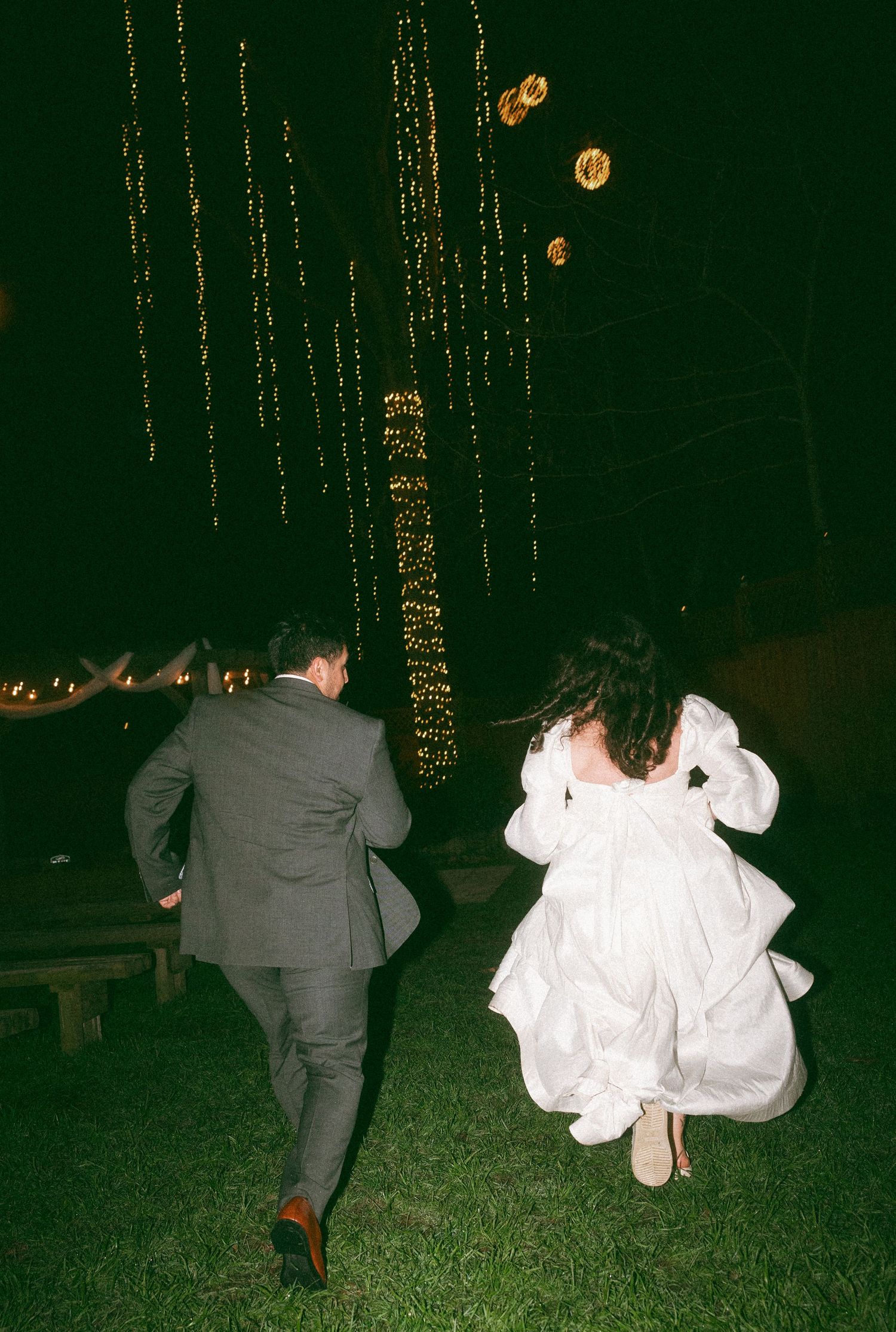 Bride and groom running across green grass at night, surrounded by twinkling fairy lights at an outdoor wedding reception.