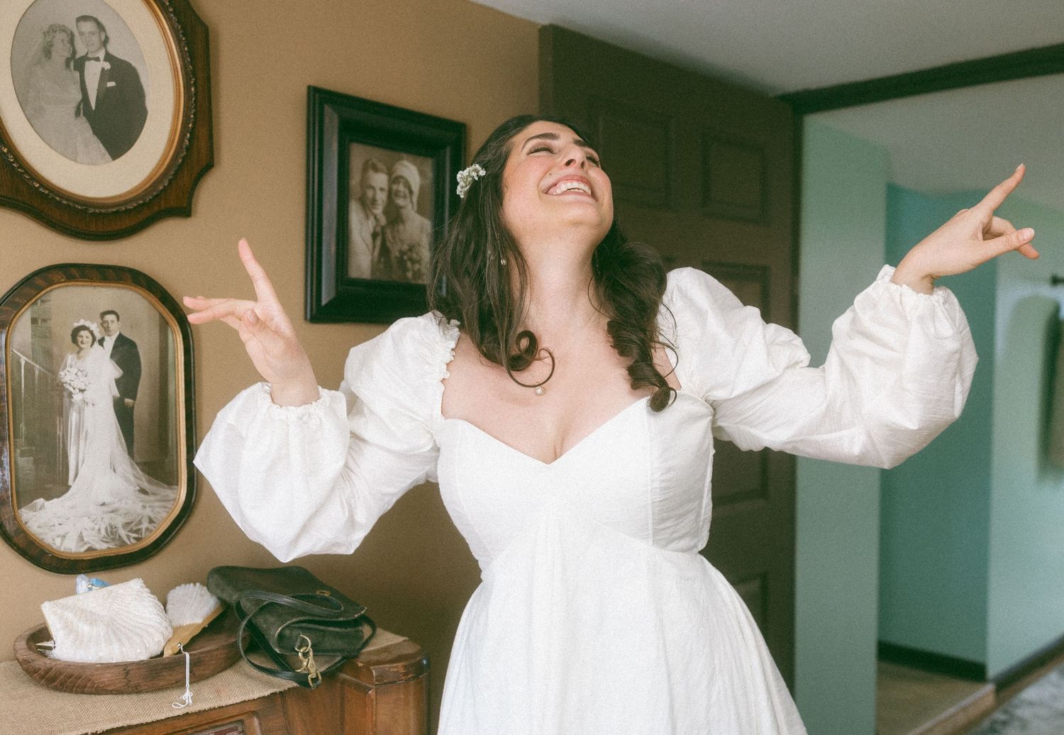 Woman in white wedding dress with puffed sleeves poses joyfully with arms outstretched in a room with vintage family photos.