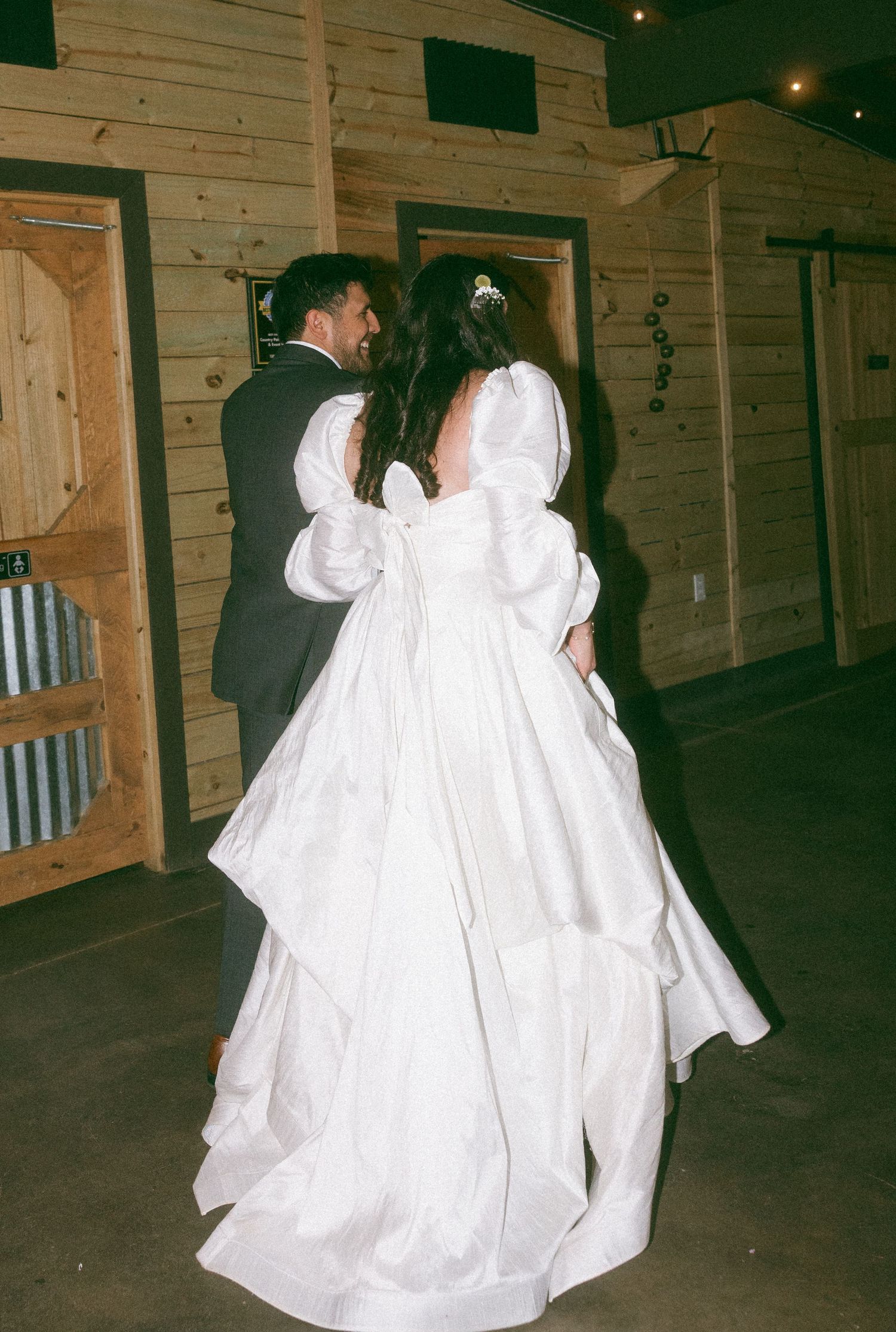 Bride in white puff-sleeve ball gown dancing with groom in black suit at rustic wood-paneled wedding venue.