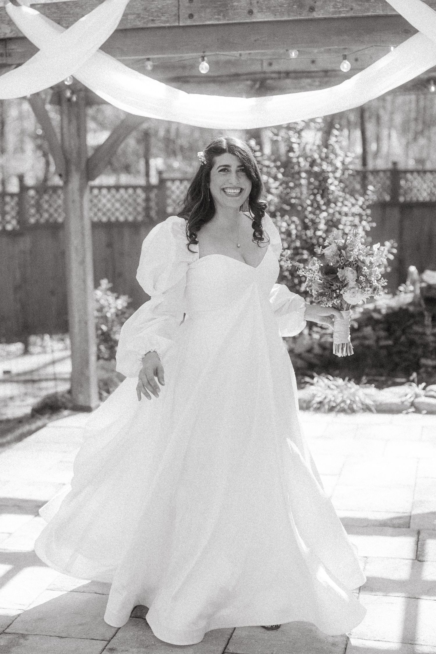 Smiling bride in a flowing white gown with puff sleeves holds a bouquet under a wooden arch.