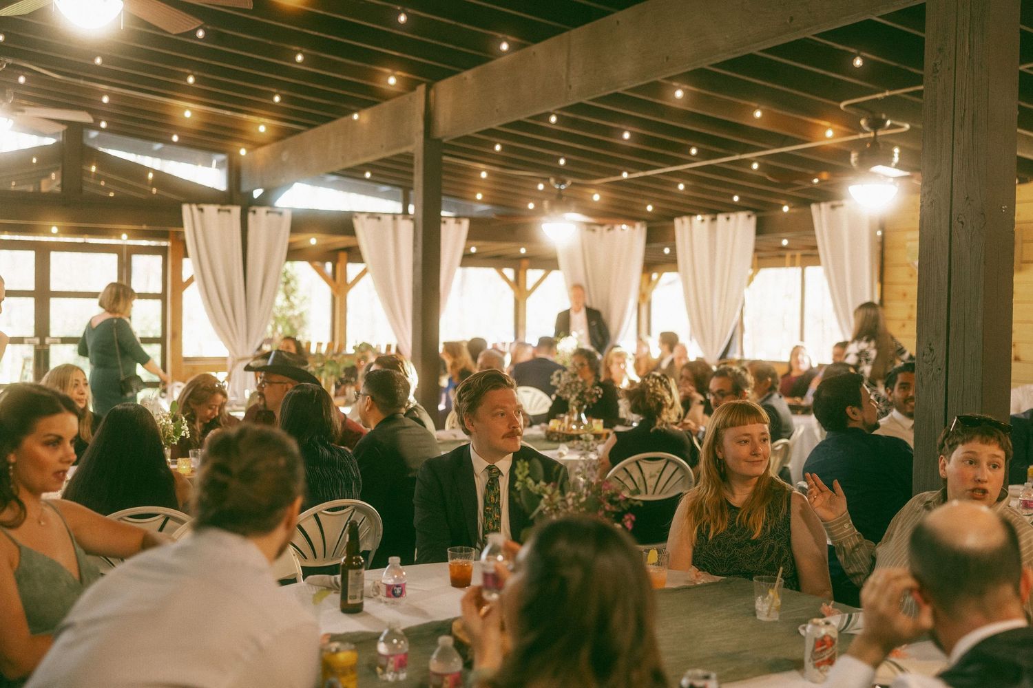 Guests seated at round tables enjoying a wedding reception dinner in a rustic barn venue with string lights.