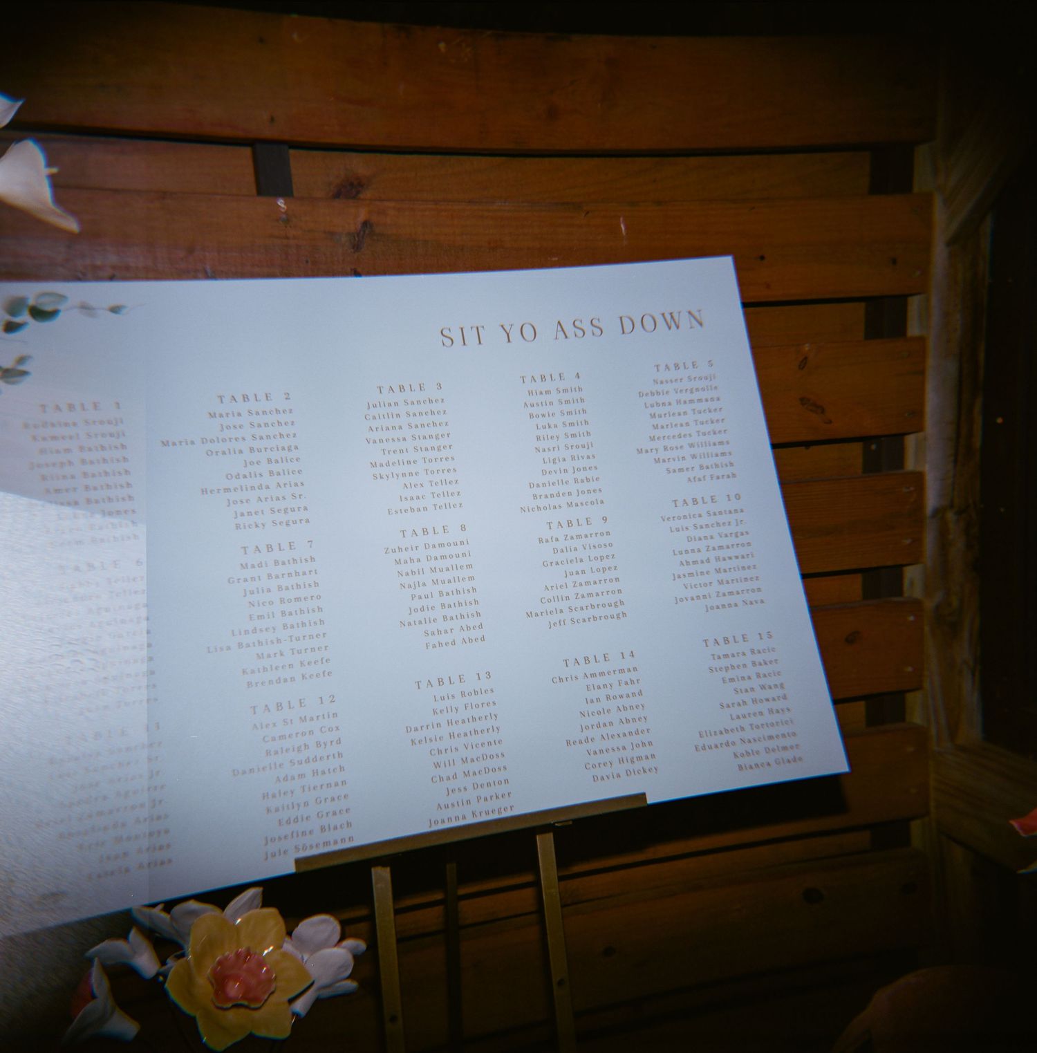 Humorous wedding seating chart sign reading Sit Yo Ass Down displayed on an easel at a rustic venue.