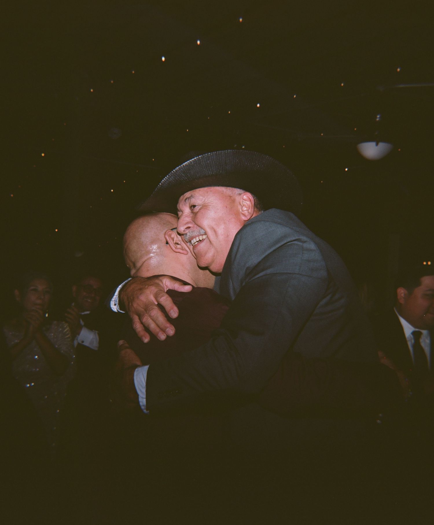 Two men sharing a warm embrace at a social gathering, one wearing a cowboy hat, captured in candid film photography.
