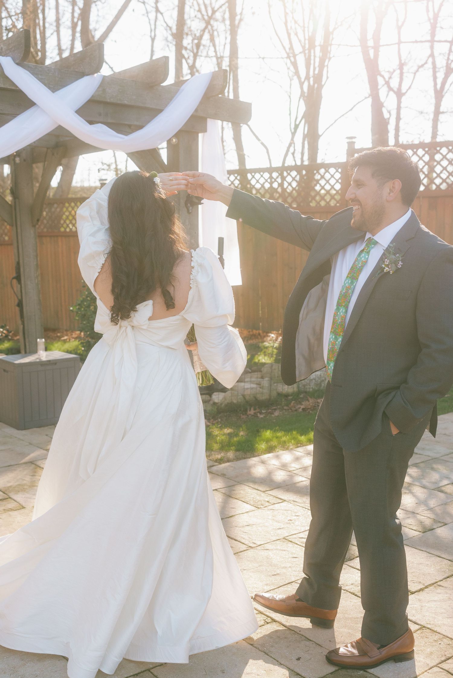 Groom twirls bride in white gown under a wooden pergola with white draping at an outdoor wedding ceremony.