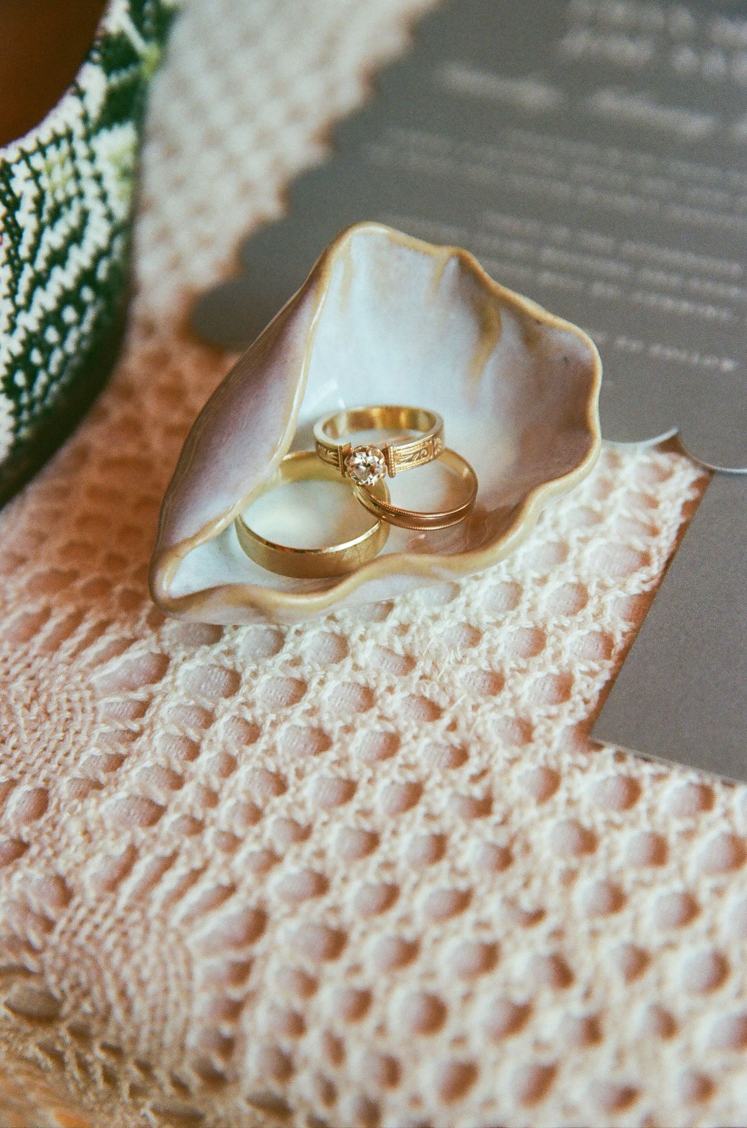 Gold wedding rings and diamond engagement ring displayed in a white shell dish on lace fabric.