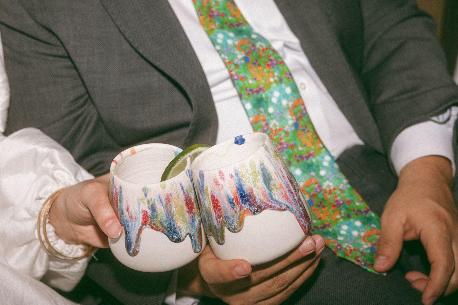 Couple holding decorative ceramic mugs with colorful painted designs, groom wearing floral tie at wedding ceremony.