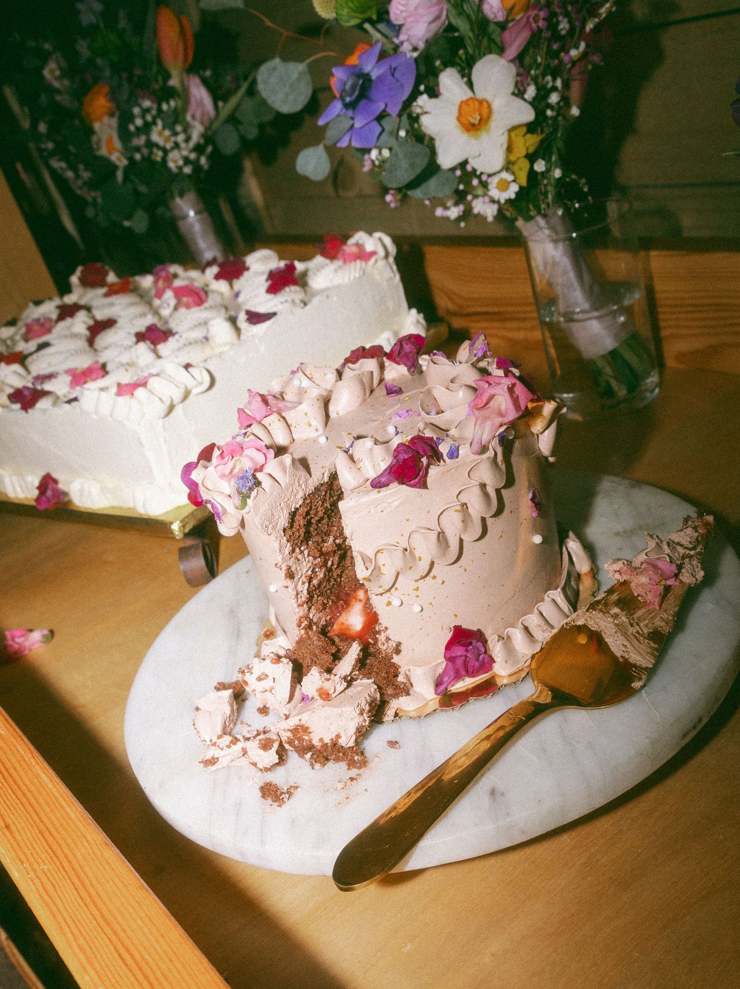 A partially sliced chocolate cake decorated with pink flowers and white frosting on a marble board.
