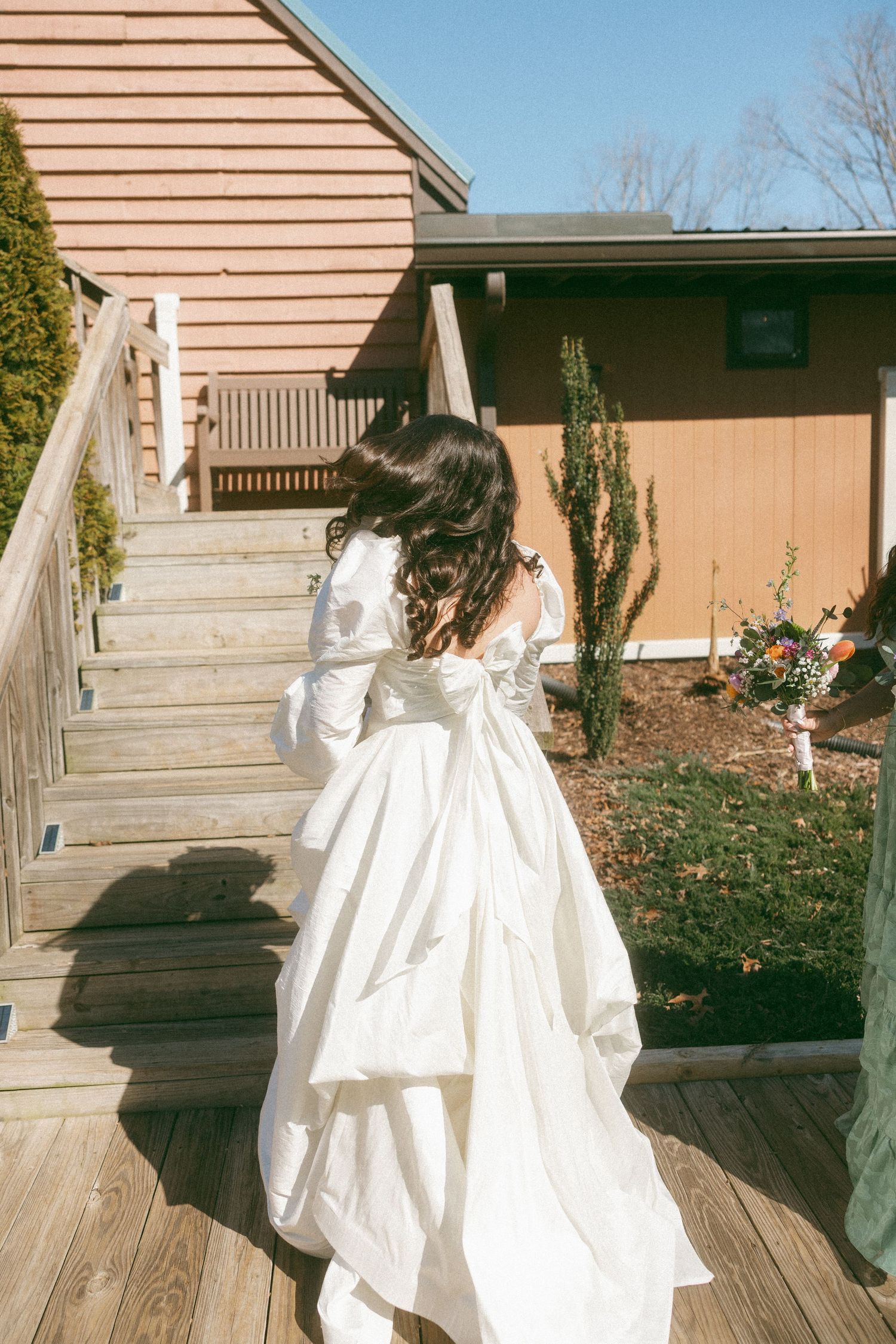 Bride in white wedding dress with bow walking up wooden deck stairs outdoors.