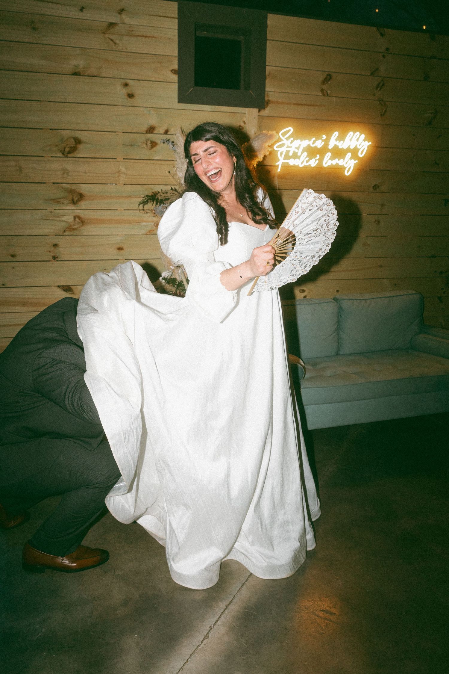 Laughing bride in white gown holding a fan, lifting her dress while dancing at a wedding reception with neon sign.
