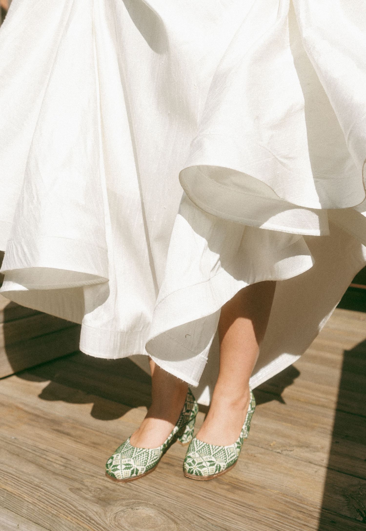 Person wearing green embroidered ballet flats beneath a flowing white dress on a wooden deck.