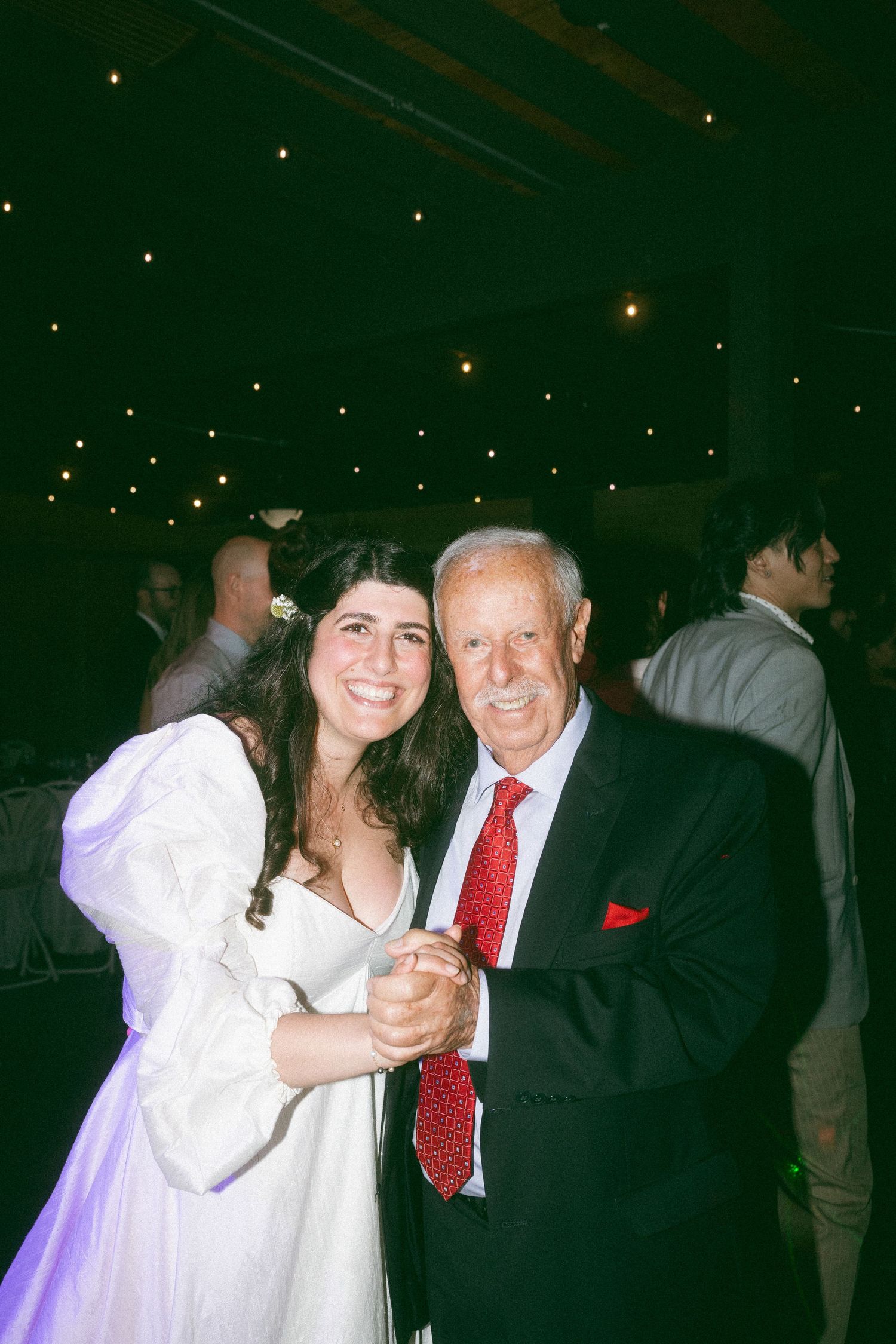 Bride in white gown dancing with elderly man in black suit and red tie at wedding reception.