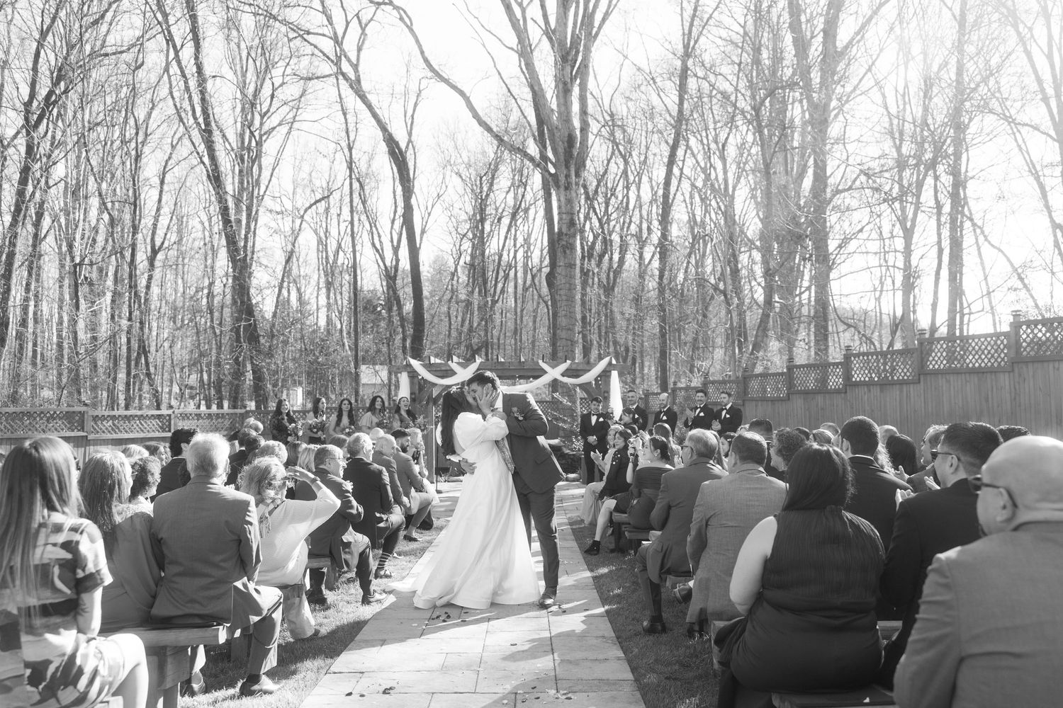 Black and white wide shot of outdoor winter wedding ceremony with guests seated among bare trees.