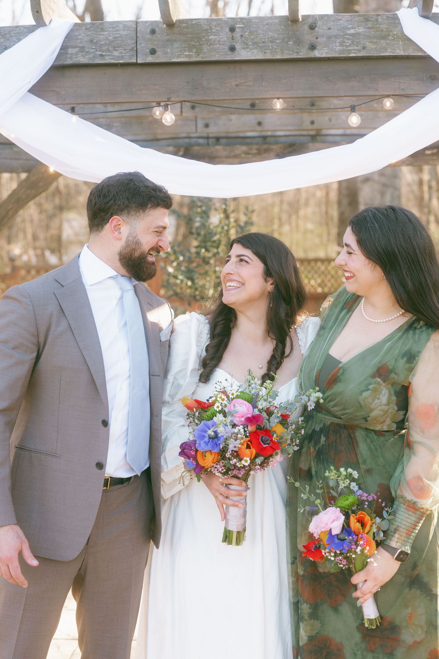Bride holding colorful bouquet smiles with groom and bridesmaid under rustic wooden arch outdoors.