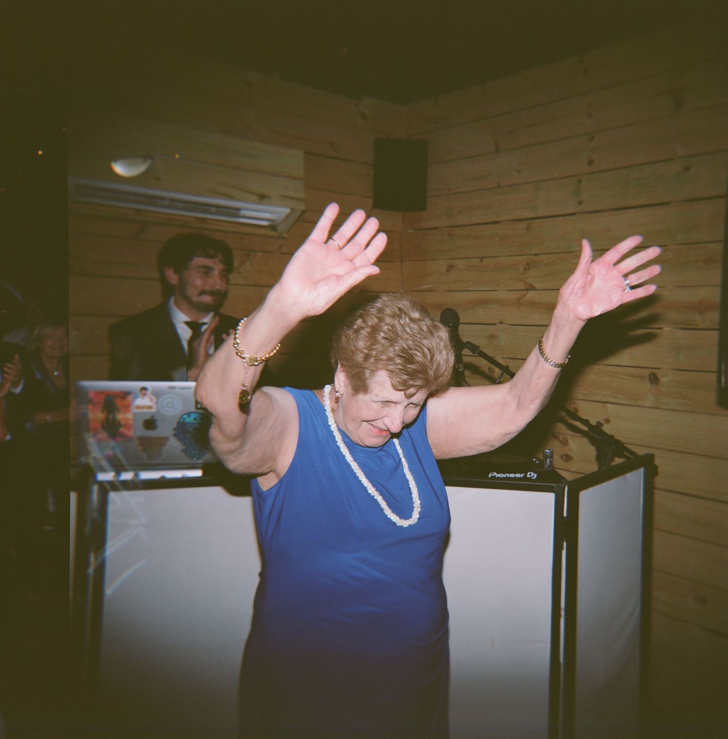 Elderly woman in blue dress dancing with arms raised at a wedding reception party.
