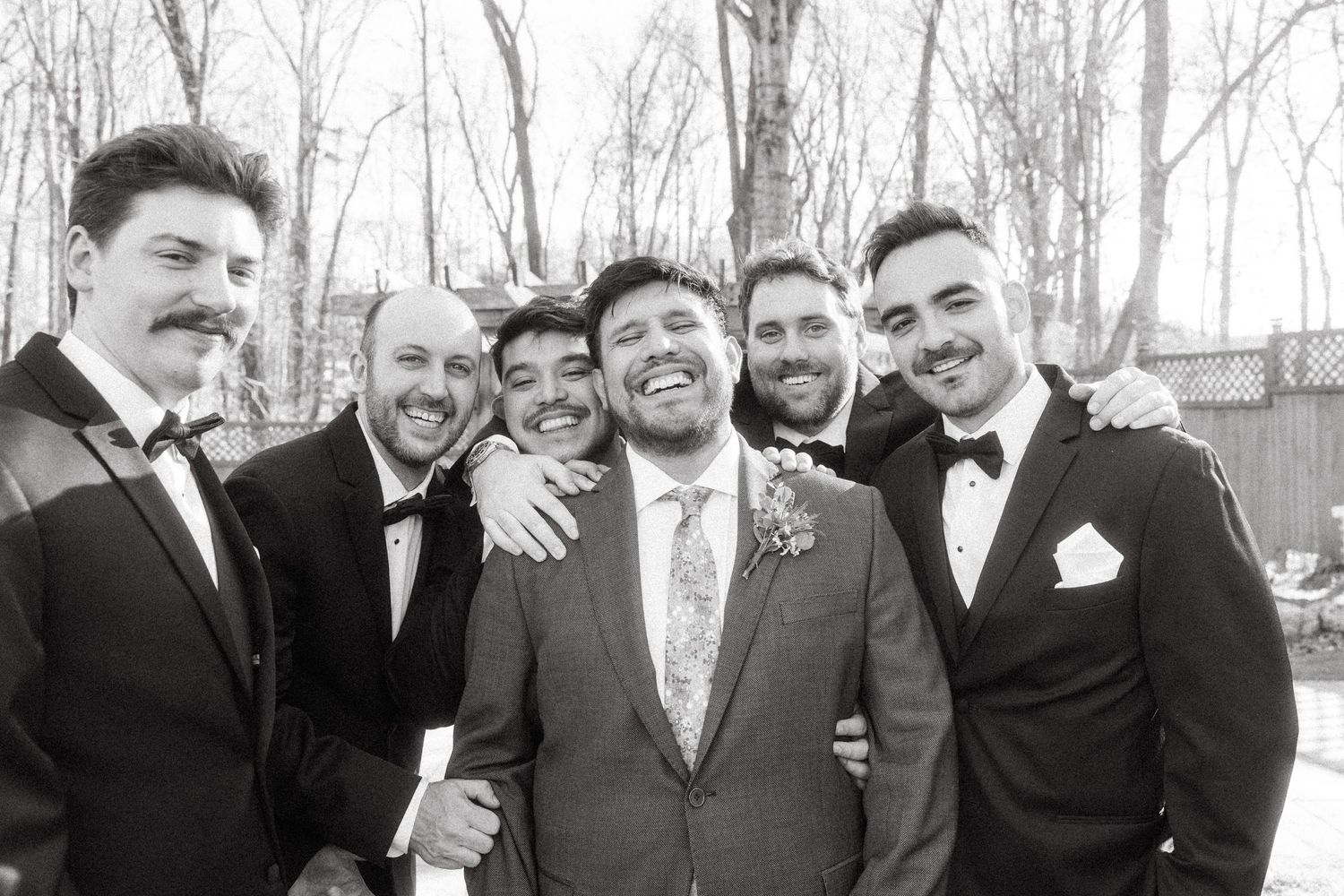 Groom smiling with five groomsmen in tuxedos in black and white outdoor wedding photo.