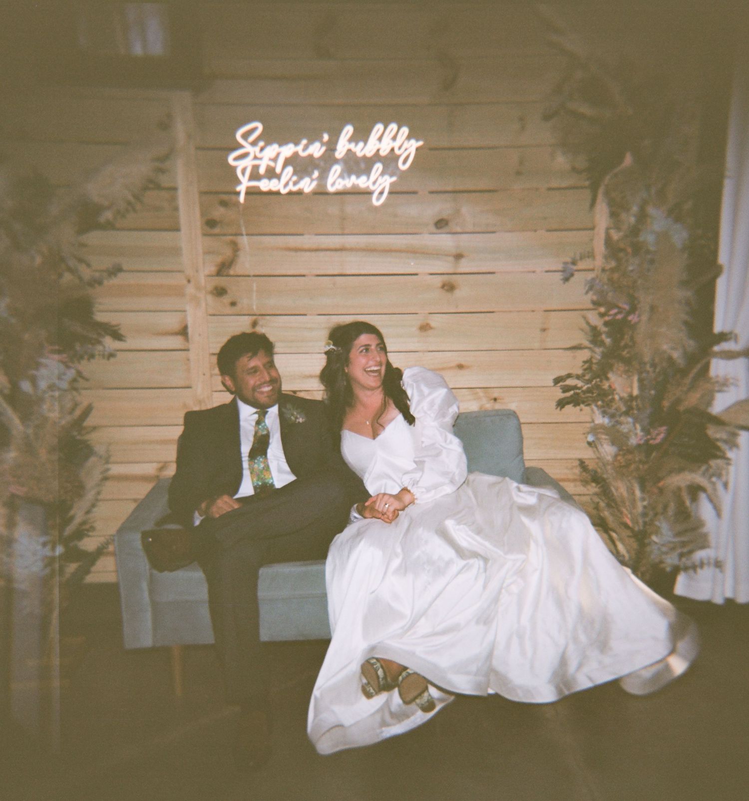 Bride and groom sitting together on a couch beneath a neon sign reading Sippin Bubbly Feelin Lovely at their wedding reception.