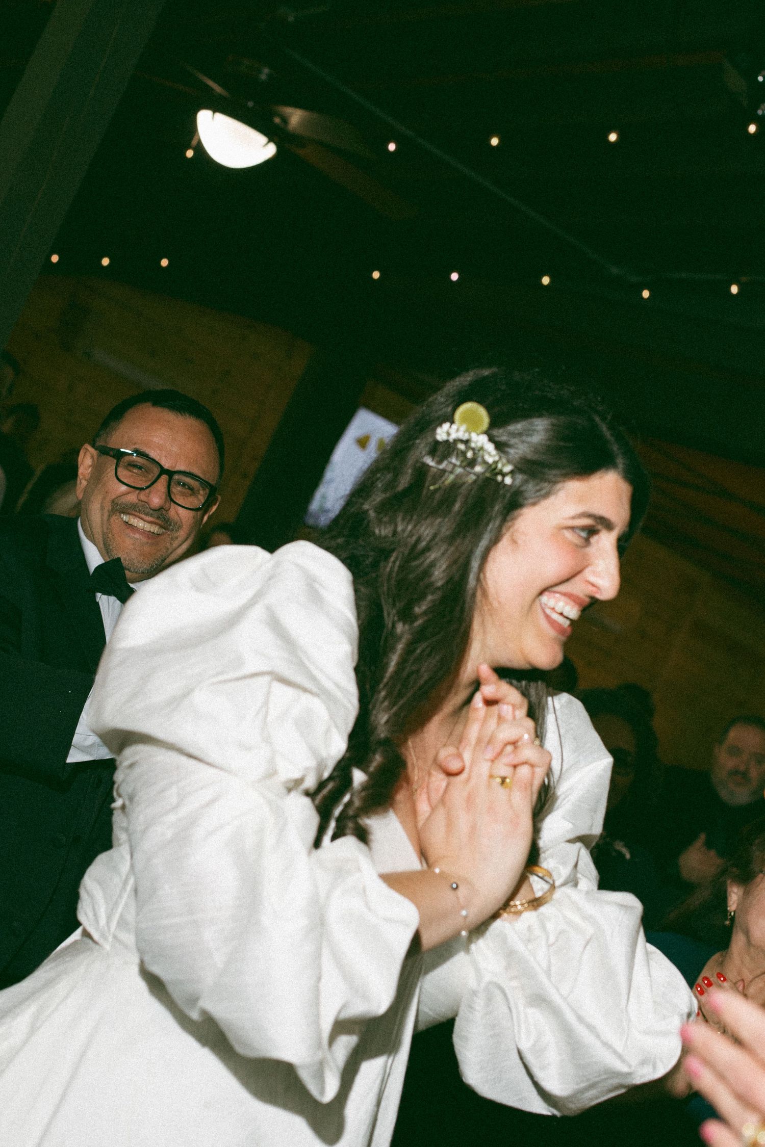 Joyful bride in white puff-sleeve wedding dress laughing and clapping at indoor wedding reception.