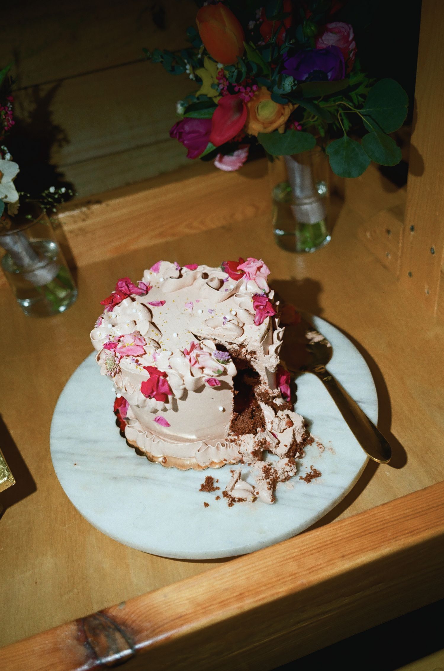 Partially sliced chocolate cake with pink floral frosting on marble plate, surrounded by colorful flower arrangements.