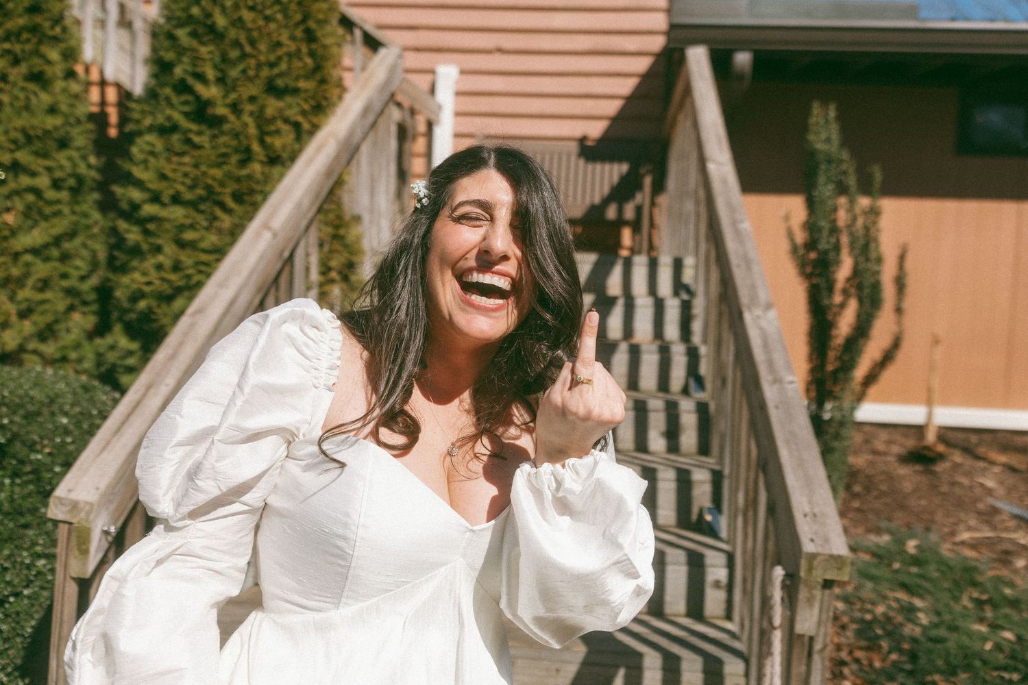 Happy bride in a white puff-sleeve dress laughing outdoors on a sunny day near wooden stairs.