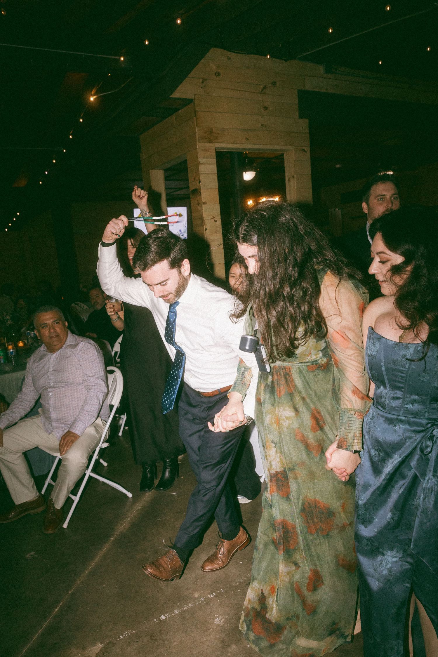 Guests dancing energetically at a rustic wedding reception venue with string lights overhead.