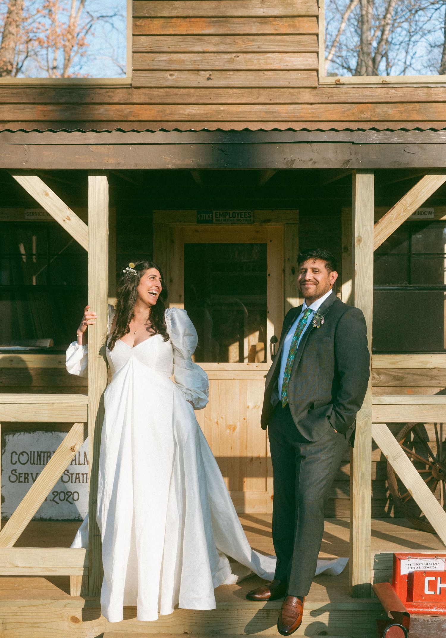 Bride and groom laughing together outside rustic wooden building on sunny winter wedding day.