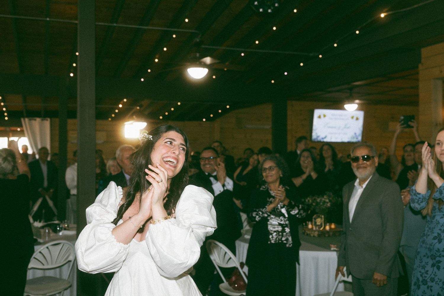 A joyful woman in a white dress claps and looks up at a celebration event in a warmly lit venue.
