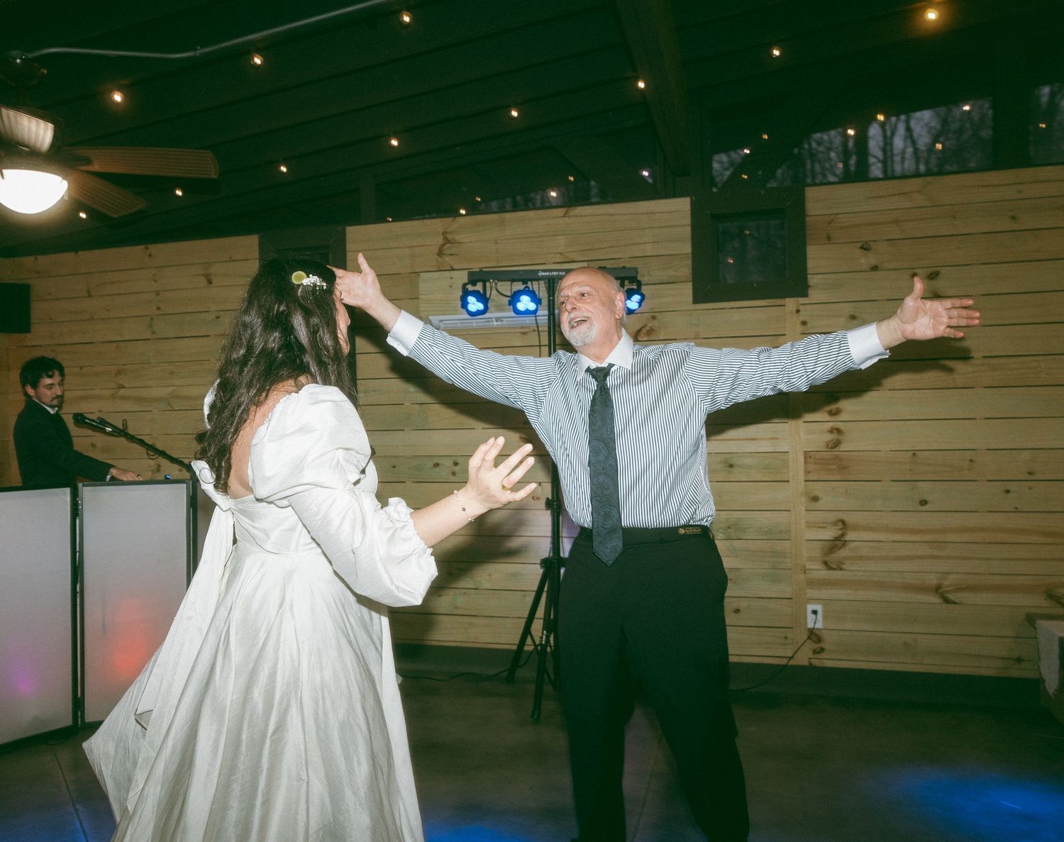 Bride in white gown and older man in suit dancing joyfully at a wedding reception in a rustic wood-paneled venue.