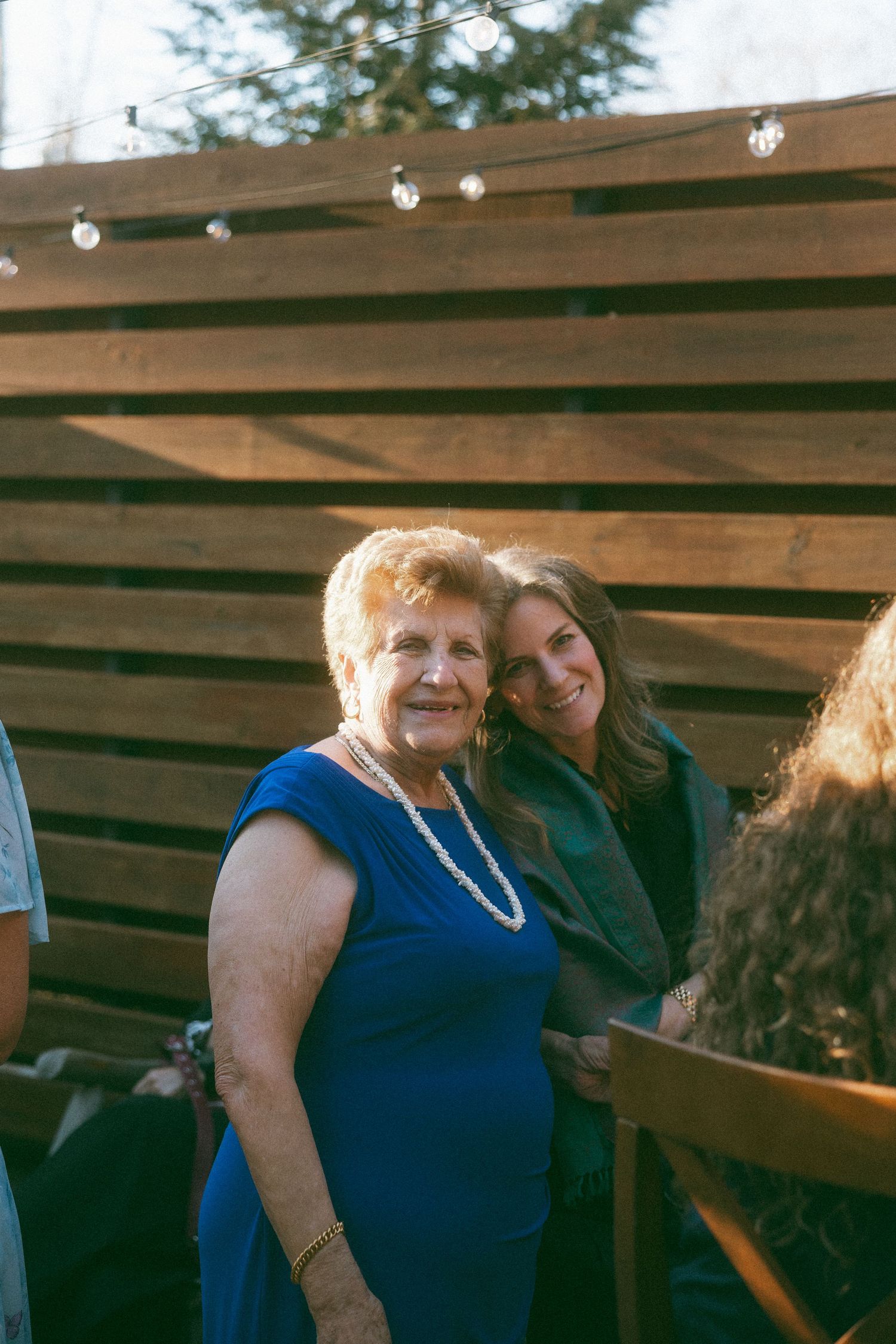 Two women smiling together at an outdoor event, one in a blue dress with pearl necklace, against a wooden fence backdrop.