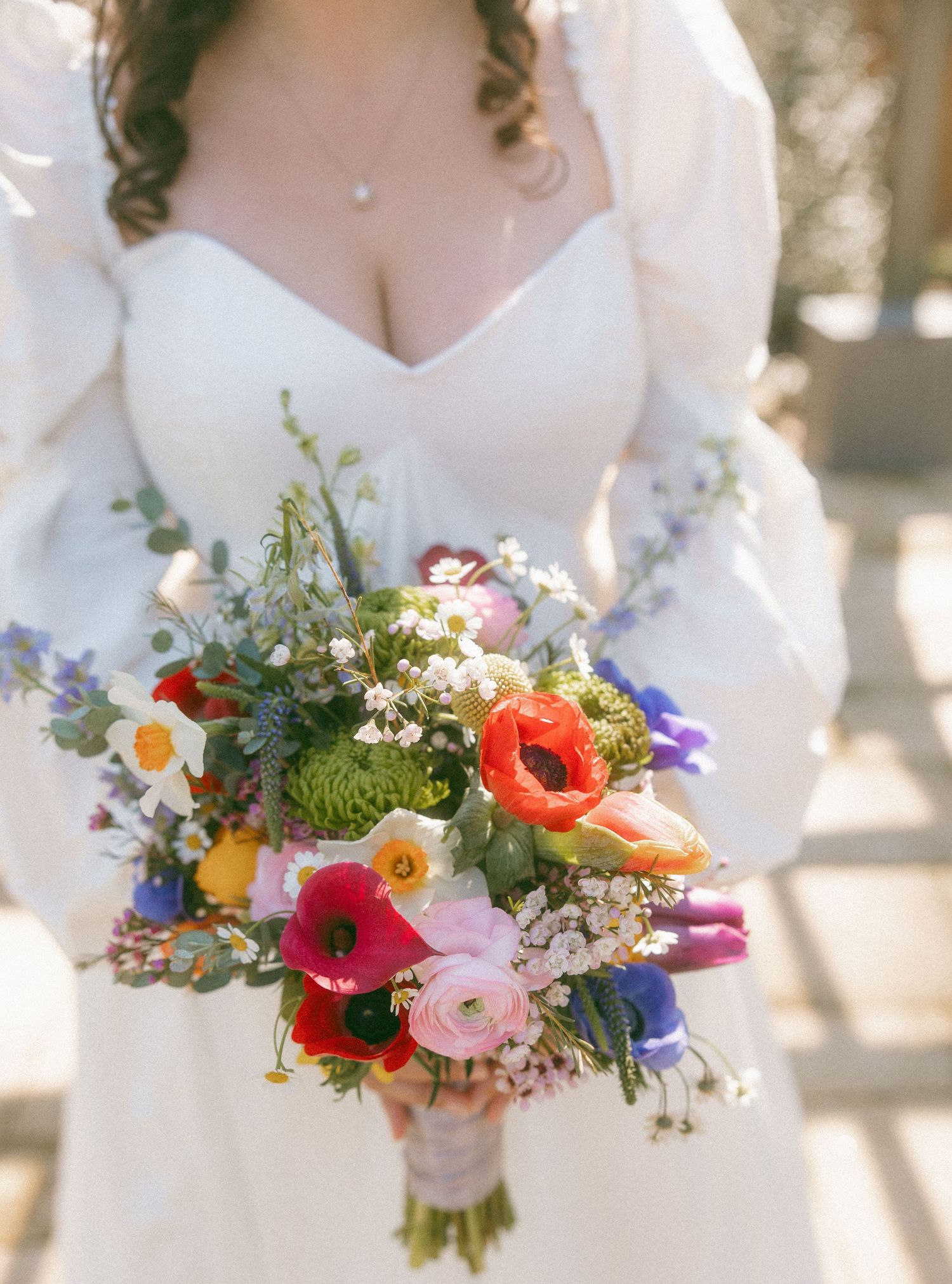 Bride in white long-sleeve gown holding a vibrant colorful bouquet of poppies, ranunculus, and wildflowers.