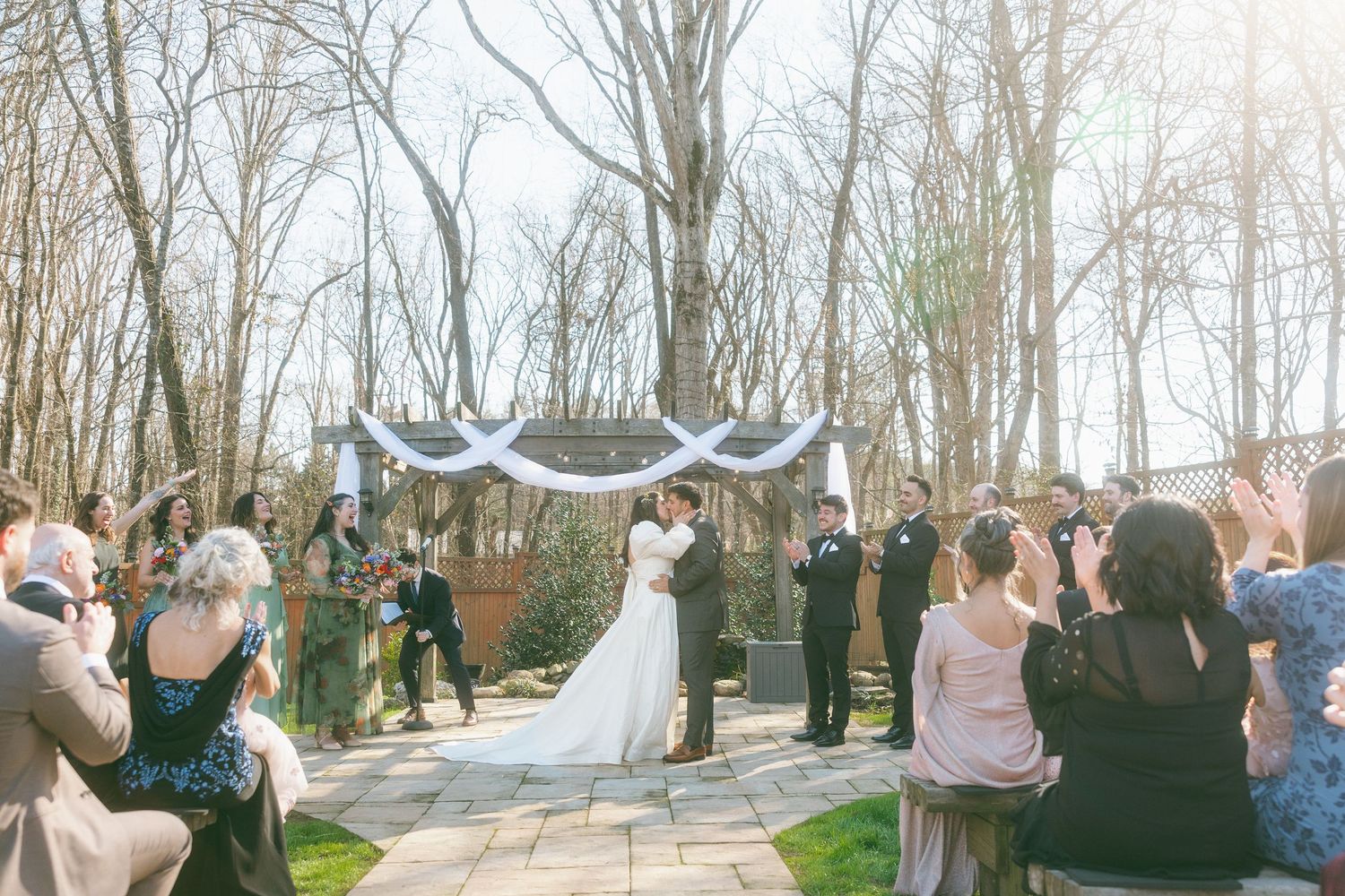 Bride and groom share a kiss at an outdoor wedding ceremony under a draped arch with seated guests.