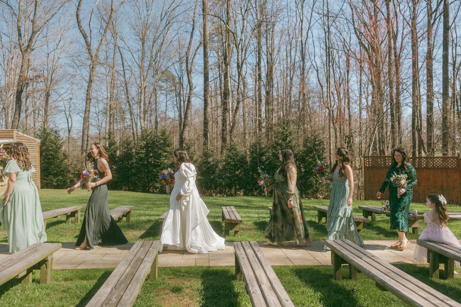 Bride in white gown walks toward bridesmaids in green and sage dresses at outdoor wedding.