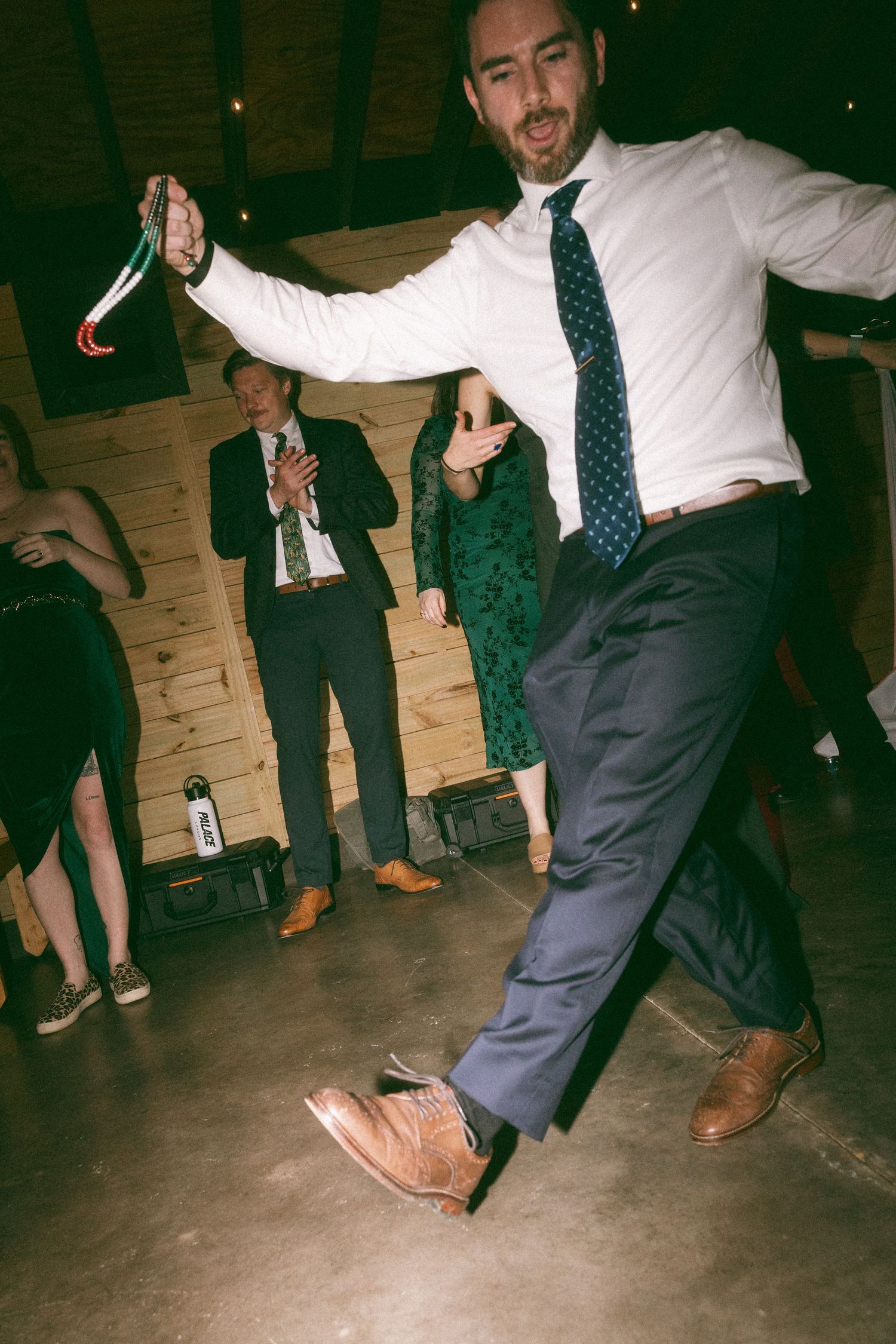 Man in white shirt and tie dancing energetically at a wedding reception in a rustic wood-paneled venue.