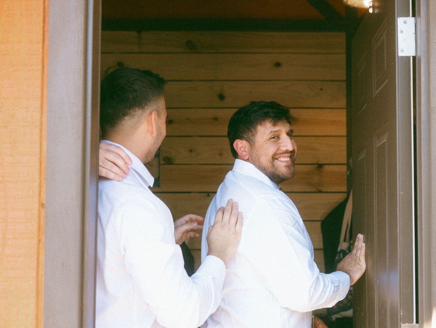 Two groomsmen in white dress shirts share a moment in a rustic wooden doorway before a wedding ceremony.