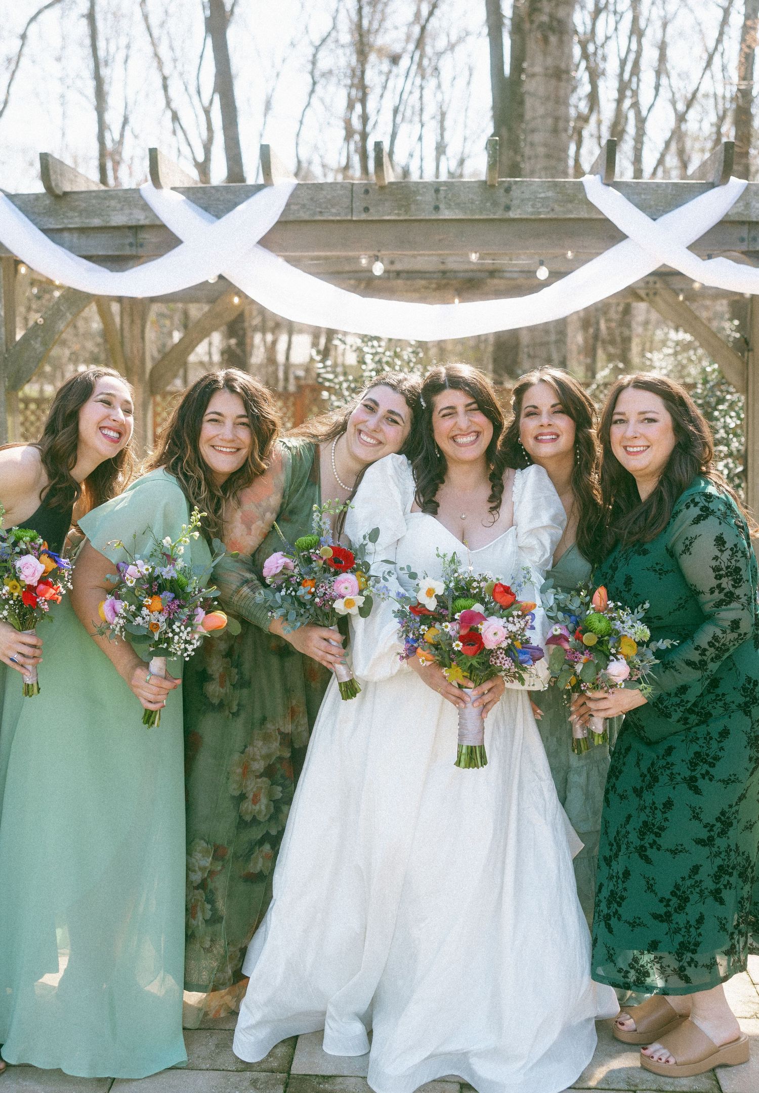 Smiling bride in white gown poses with four bridesmaids in mixed green dresses holding colorful bouquets outdoors.