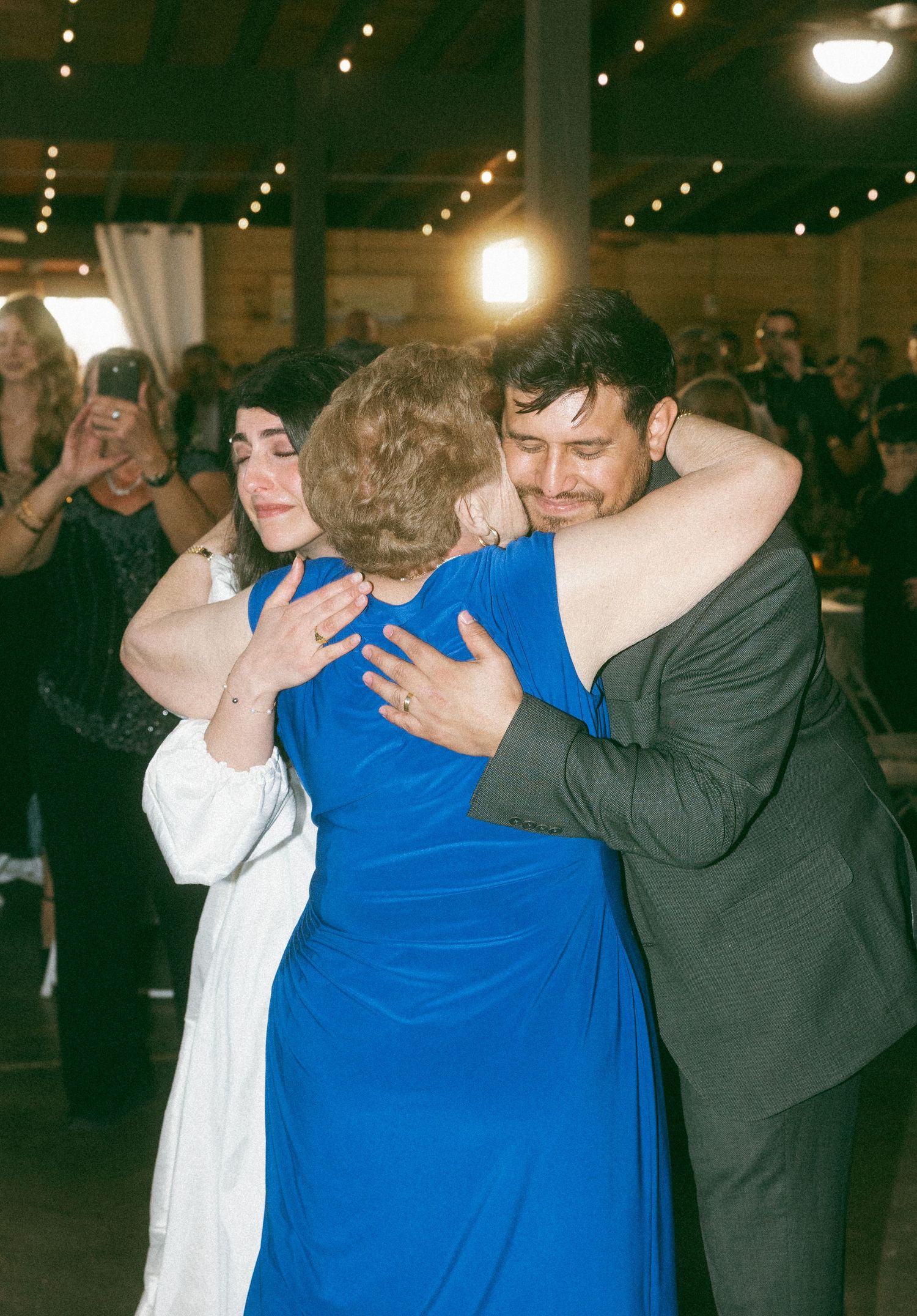 Bride, groom, and elderly woman share an emotional group hug on the dance floor at a wedding reception.