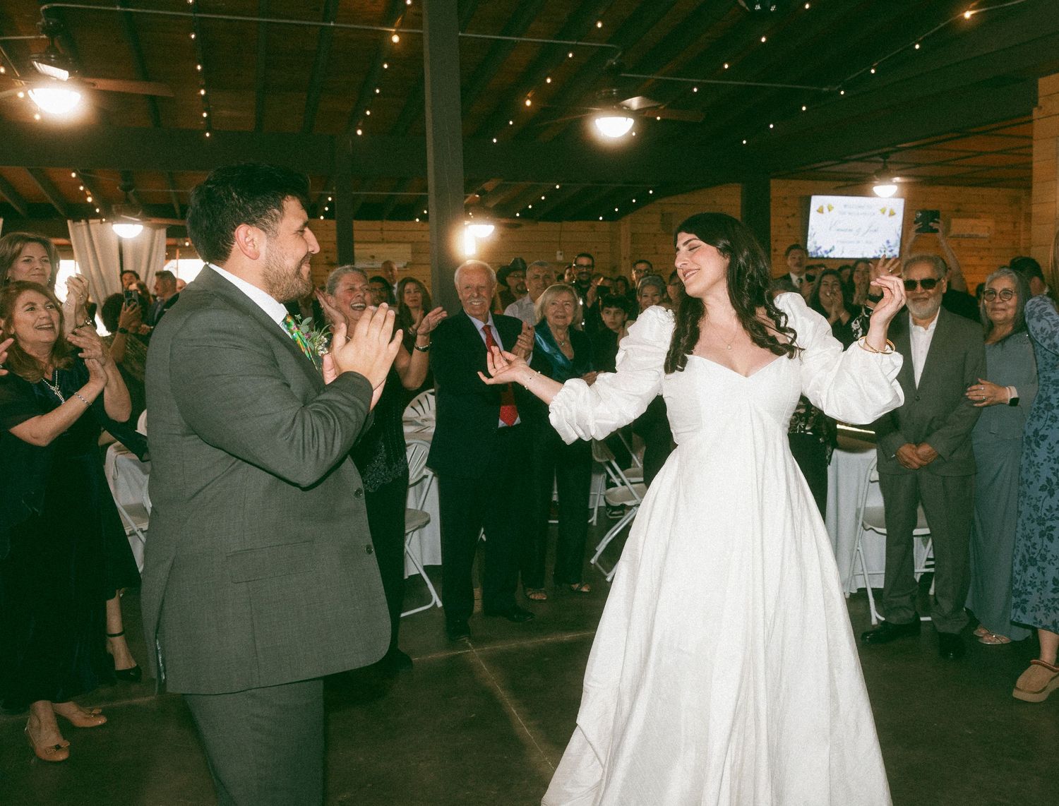 Bride in white gown and groom in suit share first dance at rustic barn wedding reception with guests applauding.