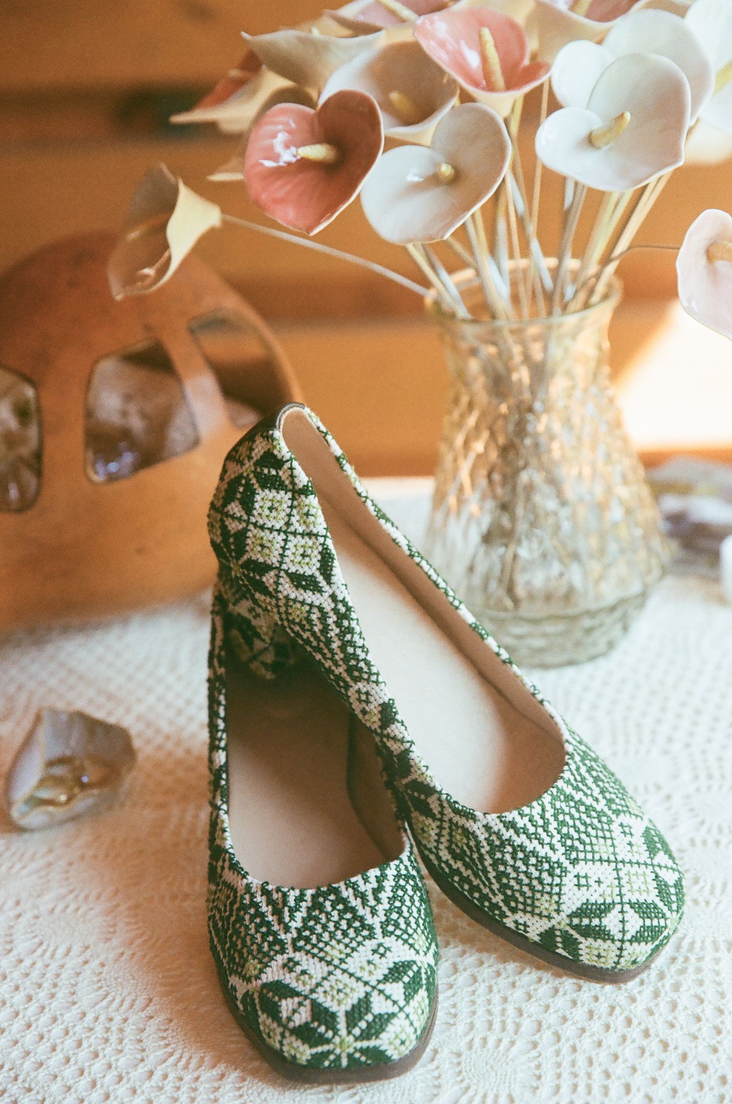 Green and white patterned ballet flats displayed on a lace surface with a decorative vase and wooden figurine.