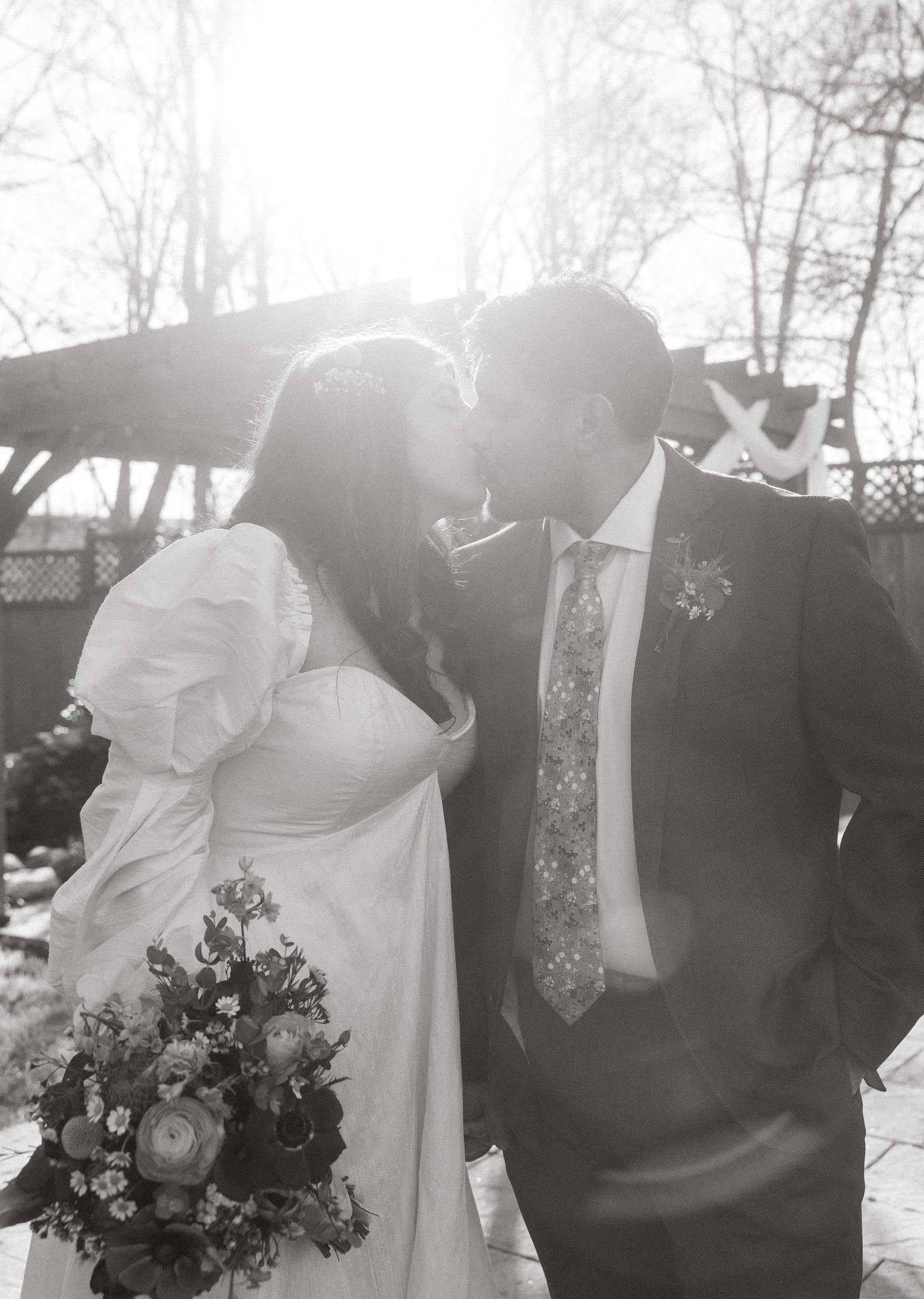 Black and white photo of bride and groom sharing a kiss backlit by sunlight holding bridal bouquet.