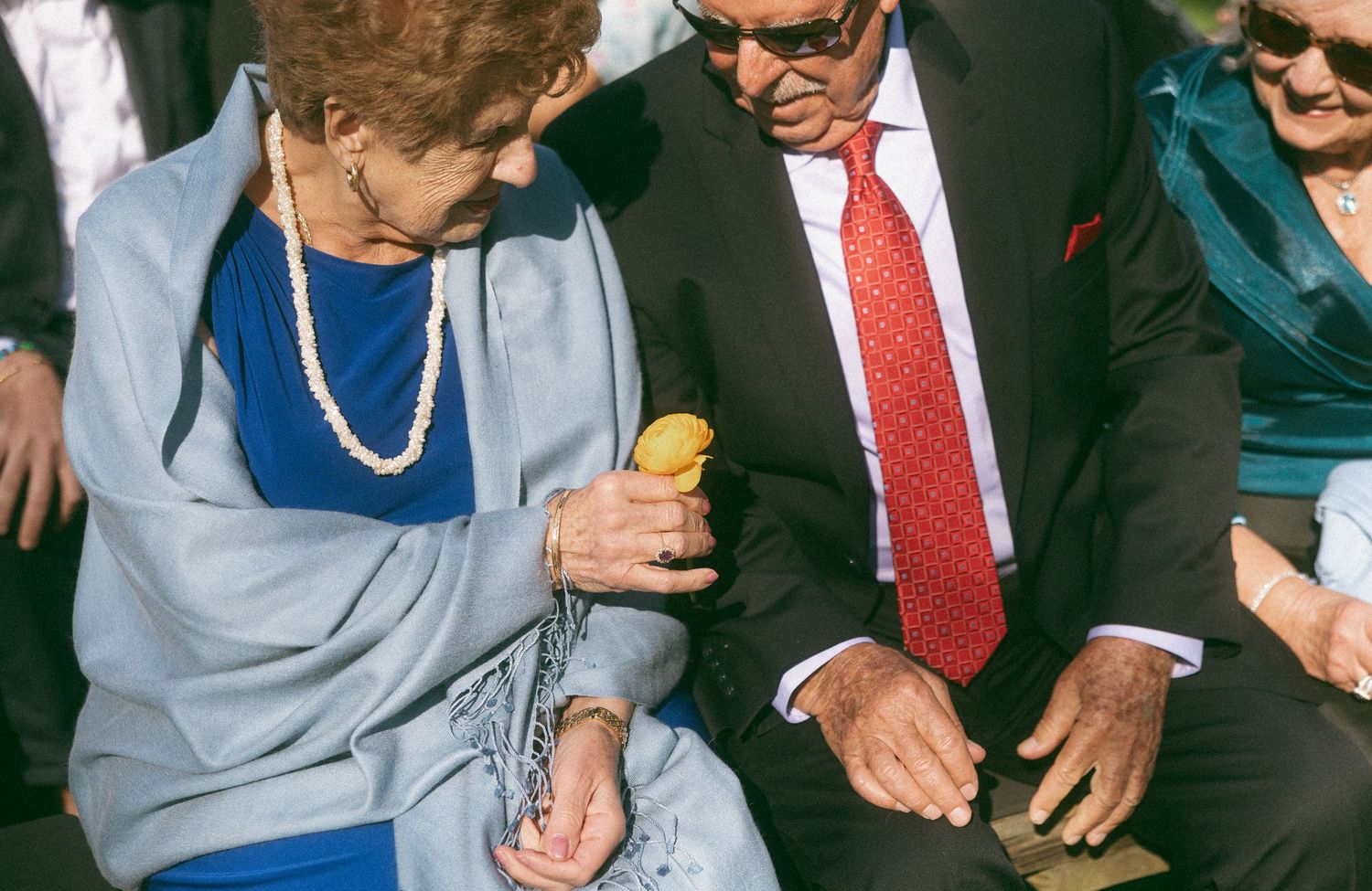Elderly couple sitting together at an outdoor wedding ceremony, woman in blue dress with pearl necklace holding a drink.