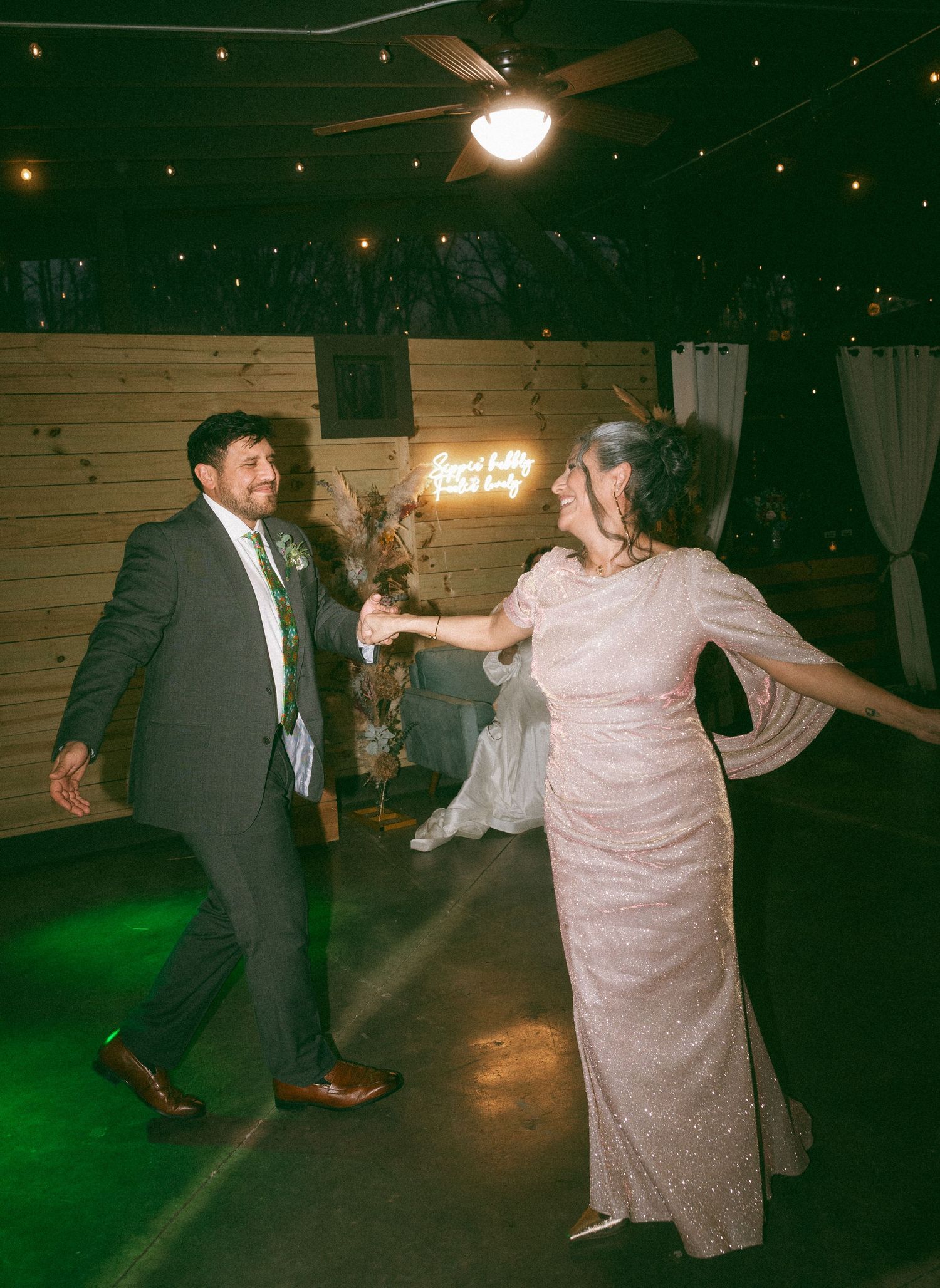 Couple dancing joyfully at a rustic barn wedding reception with neon sign and string lights in background.
