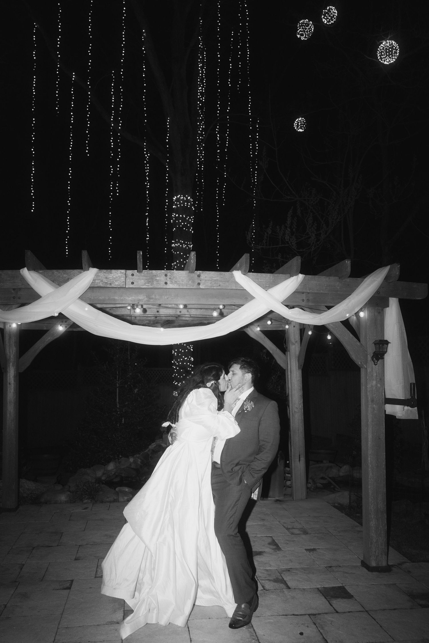 Bride and groom share a romantic kiss under a wooden pergola adorned with string lights at a night wedding reception.