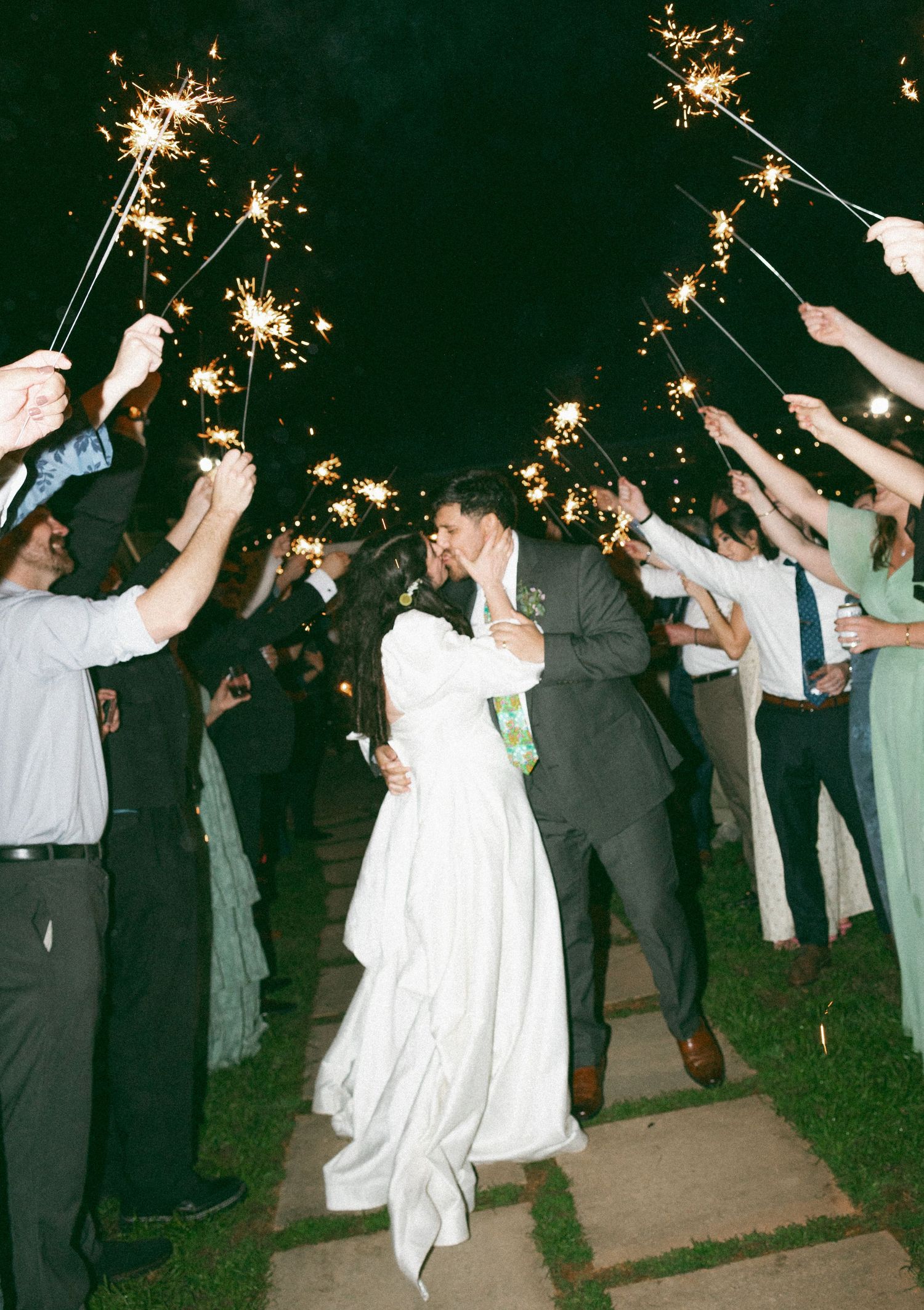 Bride and groom share a kiss during a romantic nighttime sparkler send-off surrounded by cheering wedding guests.