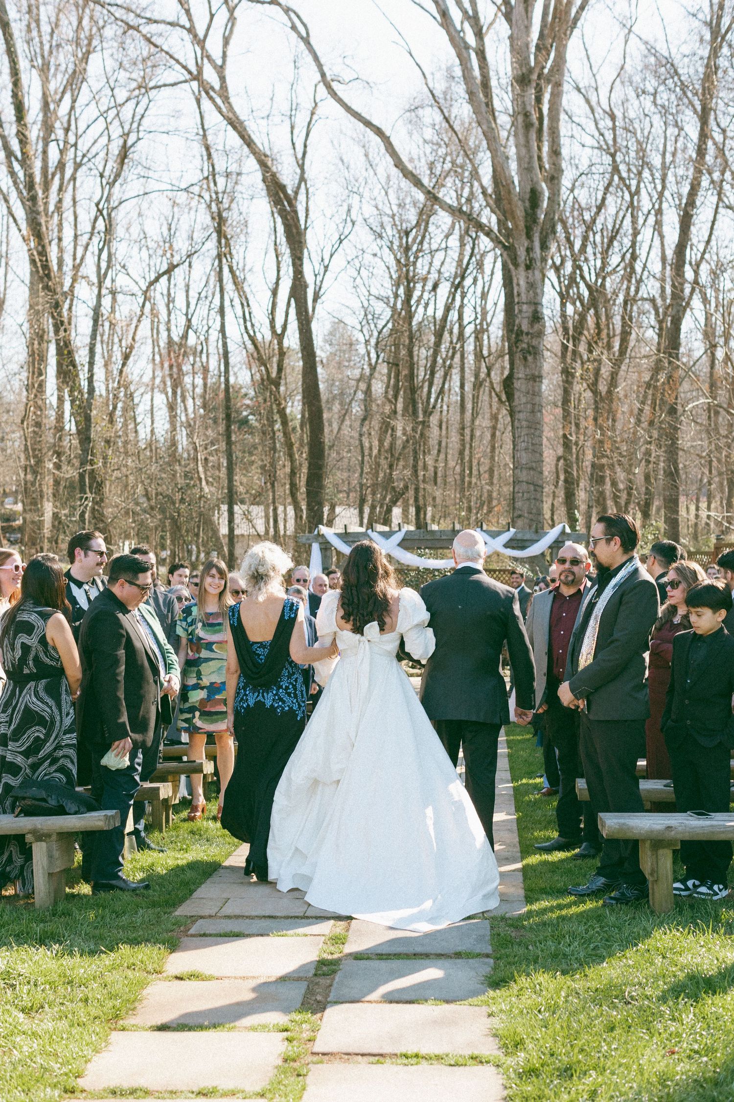Bride walking down the aisle with escort at an outdoor wedding ceremony surrounded by guests.